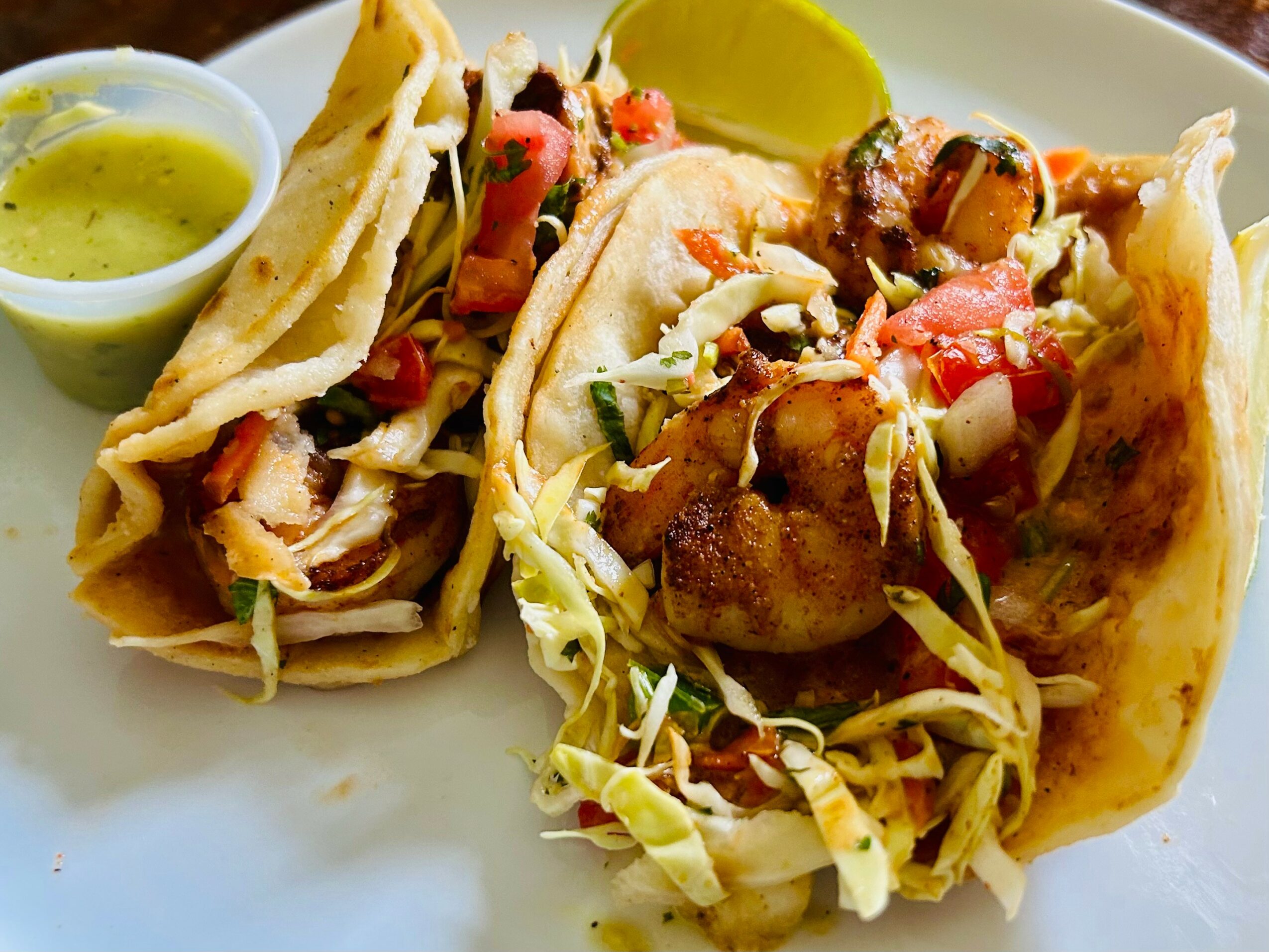 Two tacos filled with meat, vegetables, and shredded lettuce, served with lime wedge and green sauce on side.