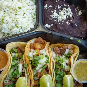 Three tacos filled with meat, topped with chopped onions and cilantro, with lime wedges and sauces nearby, rice and beans in background.