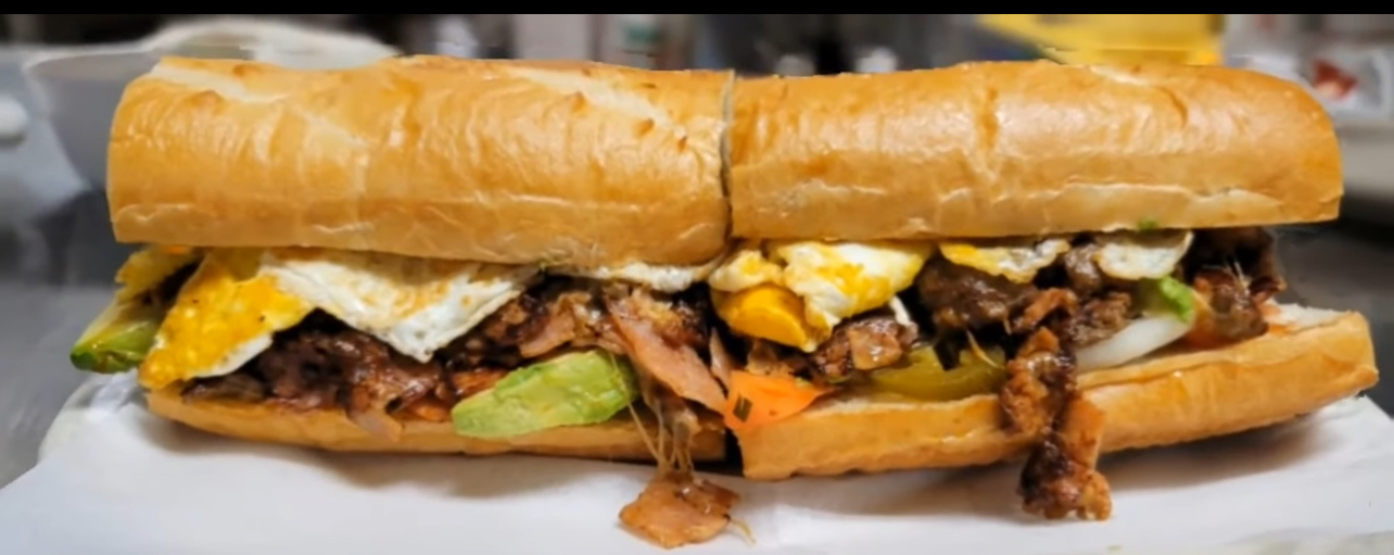 Long sandwich with meat, eggs, and vegetables in a baguette, on a white plate.