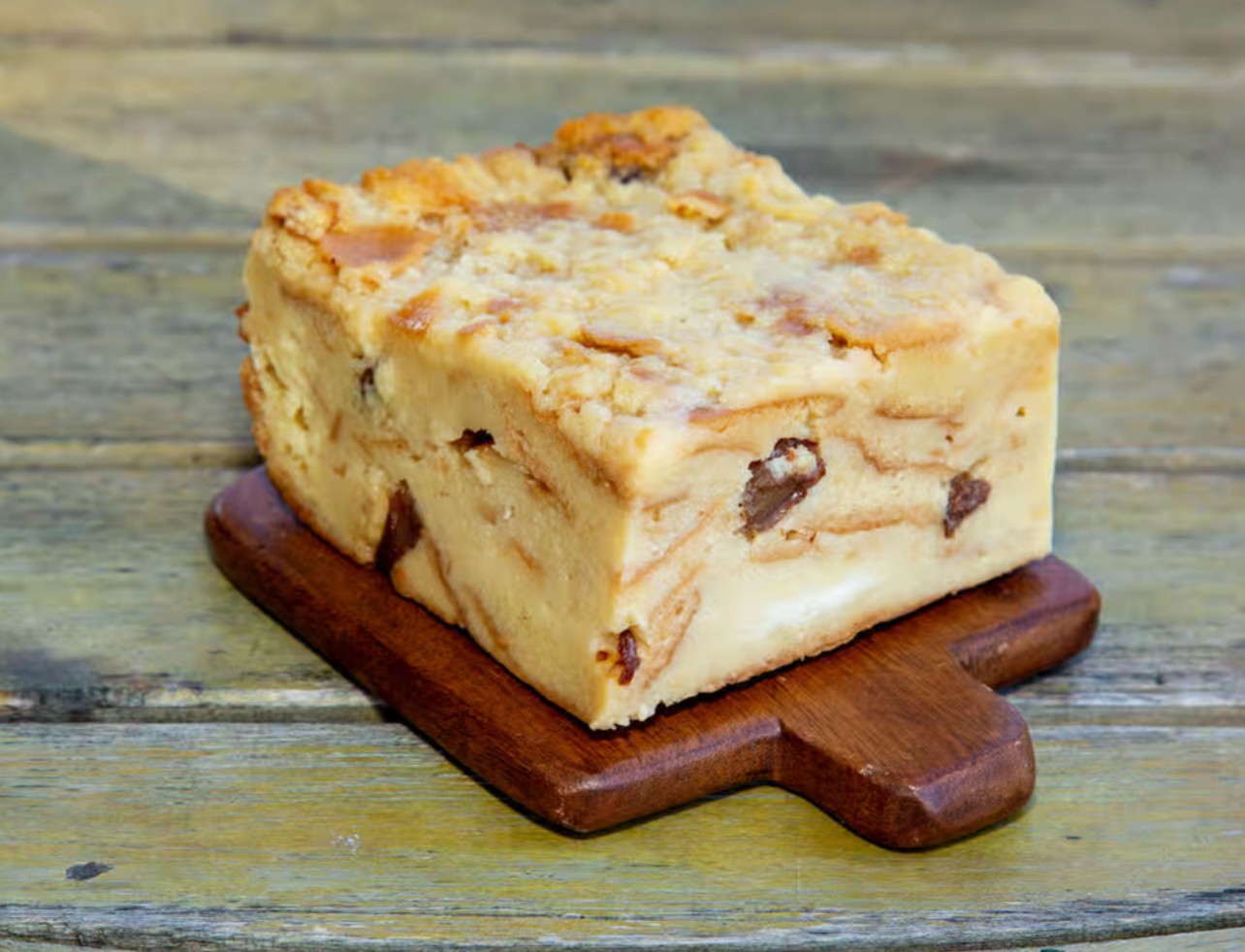 A square piece of dessert with nuts and chocolate chips on a small wooden board, on a wooden surface.