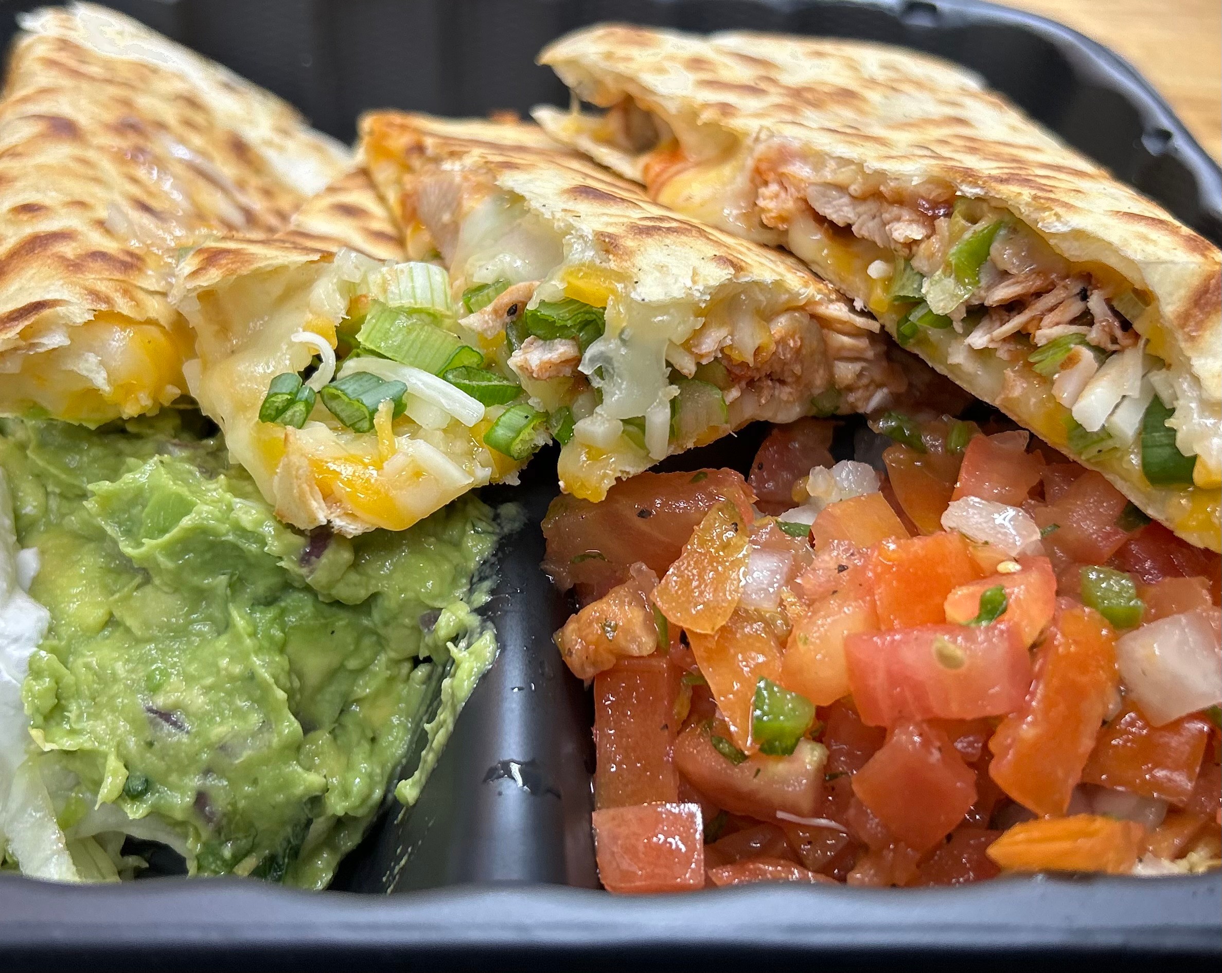 Quesadilla with melted cheese, green onions, and meat, served with guacamole and a tomato-based salsa, on a black tray.