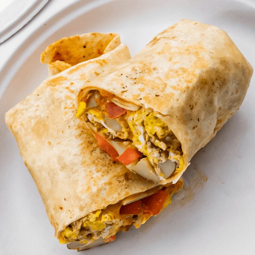 Wrapped breakfast burrito cut in half, showing scrambled eggs, tomatoes, and other ingredients inside, on a white plate.