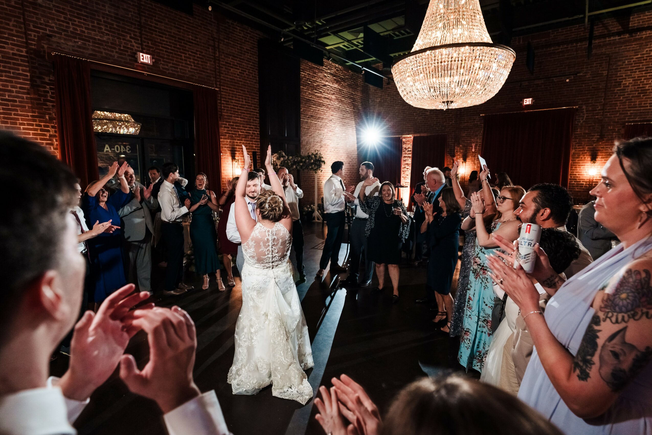 People dancing and celebrating in a dimly lit venue with a chandelier, brick walls, and a large group of guests.