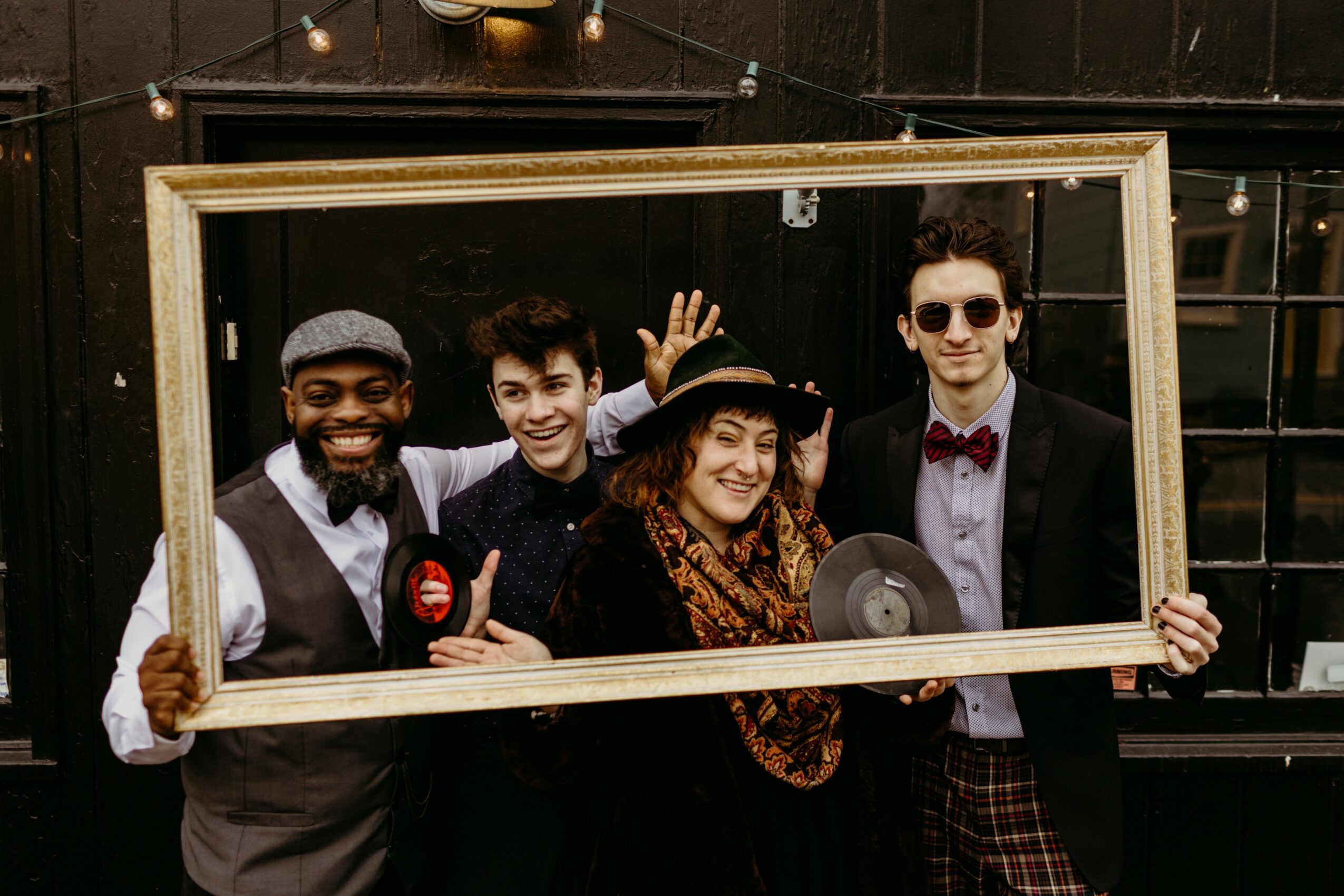 Four people holding a large empty picture frame, smiling and posing outdoors, dressed in stylish clothing.