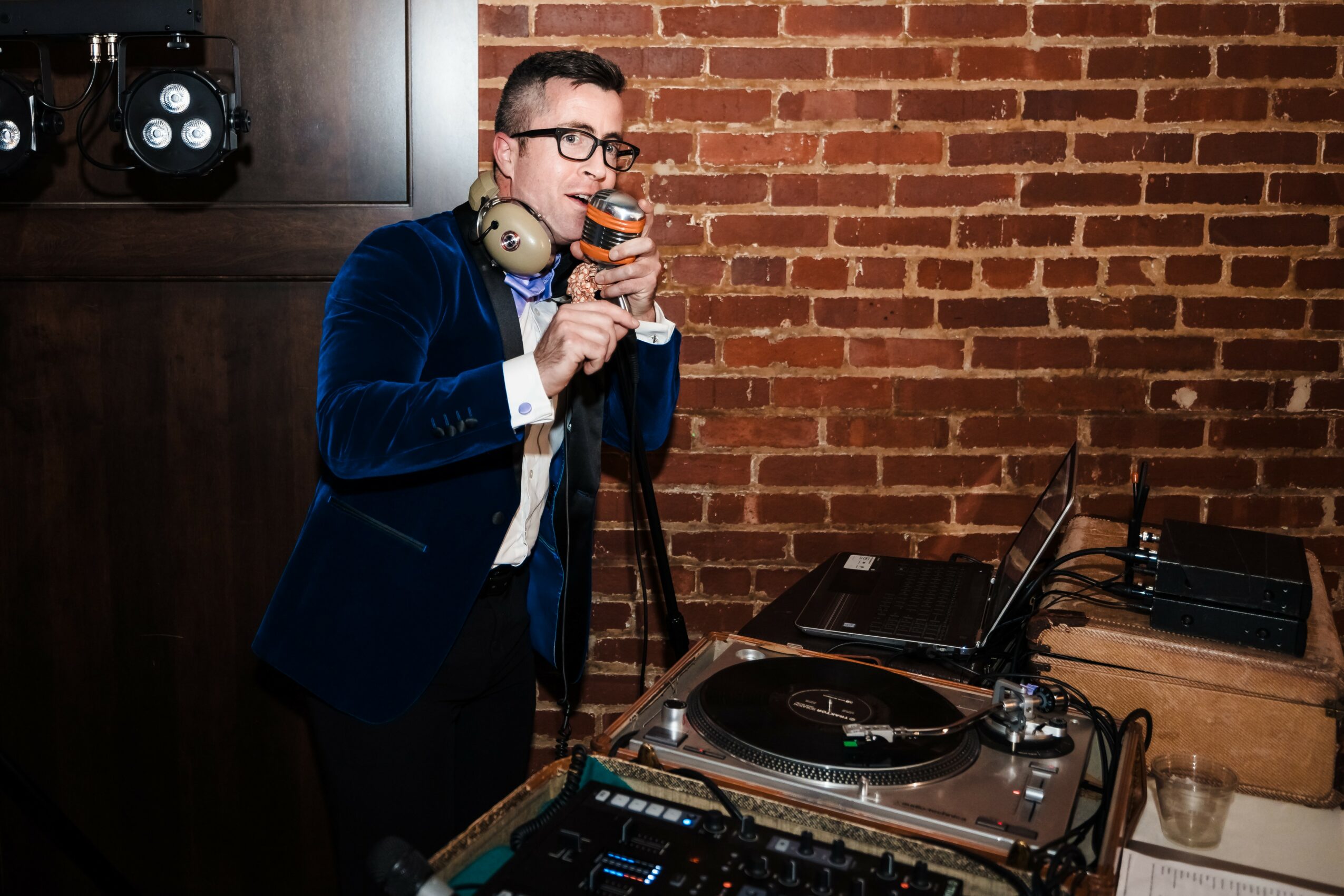 Man in glasses and blue blazer drinks from a glass, with DJ equipment and brick wall background.