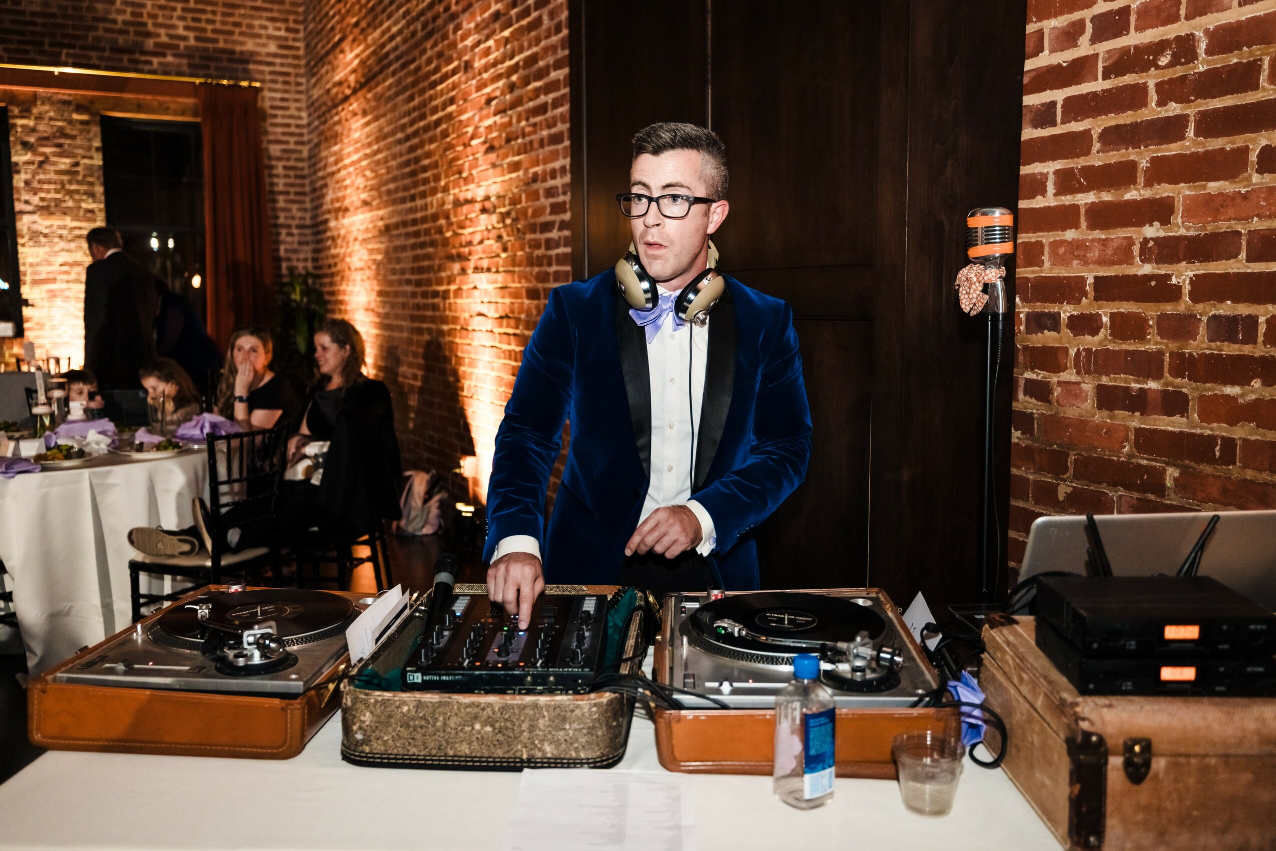 Young man in formal attire and glasses DJing at a party with guests seated at tables in the background.