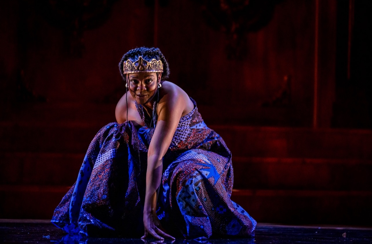 Person crouching on stage in traditional attire with a crown, dark background, and red lighting.