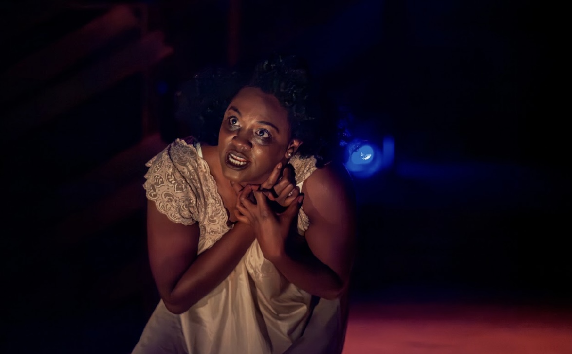 Person with curly hair and expressive face, wearing a light-colored dress, in a dark setting with blue light, hands near chest.