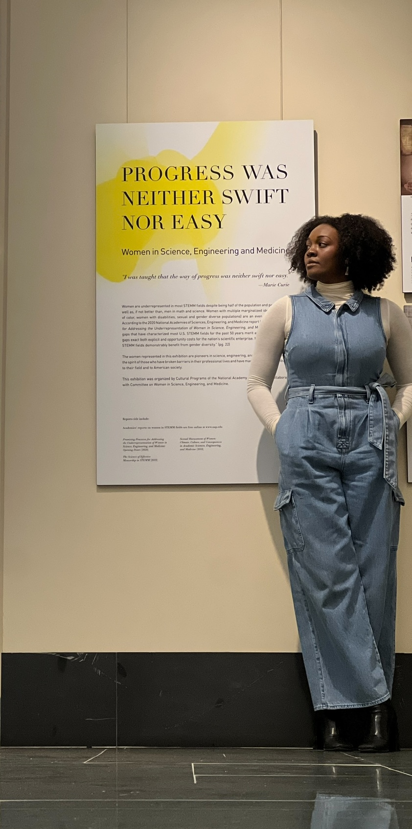 A mannequin with curly hair wearing a denim jumpsuit stands next to a large poster on a beige wall.