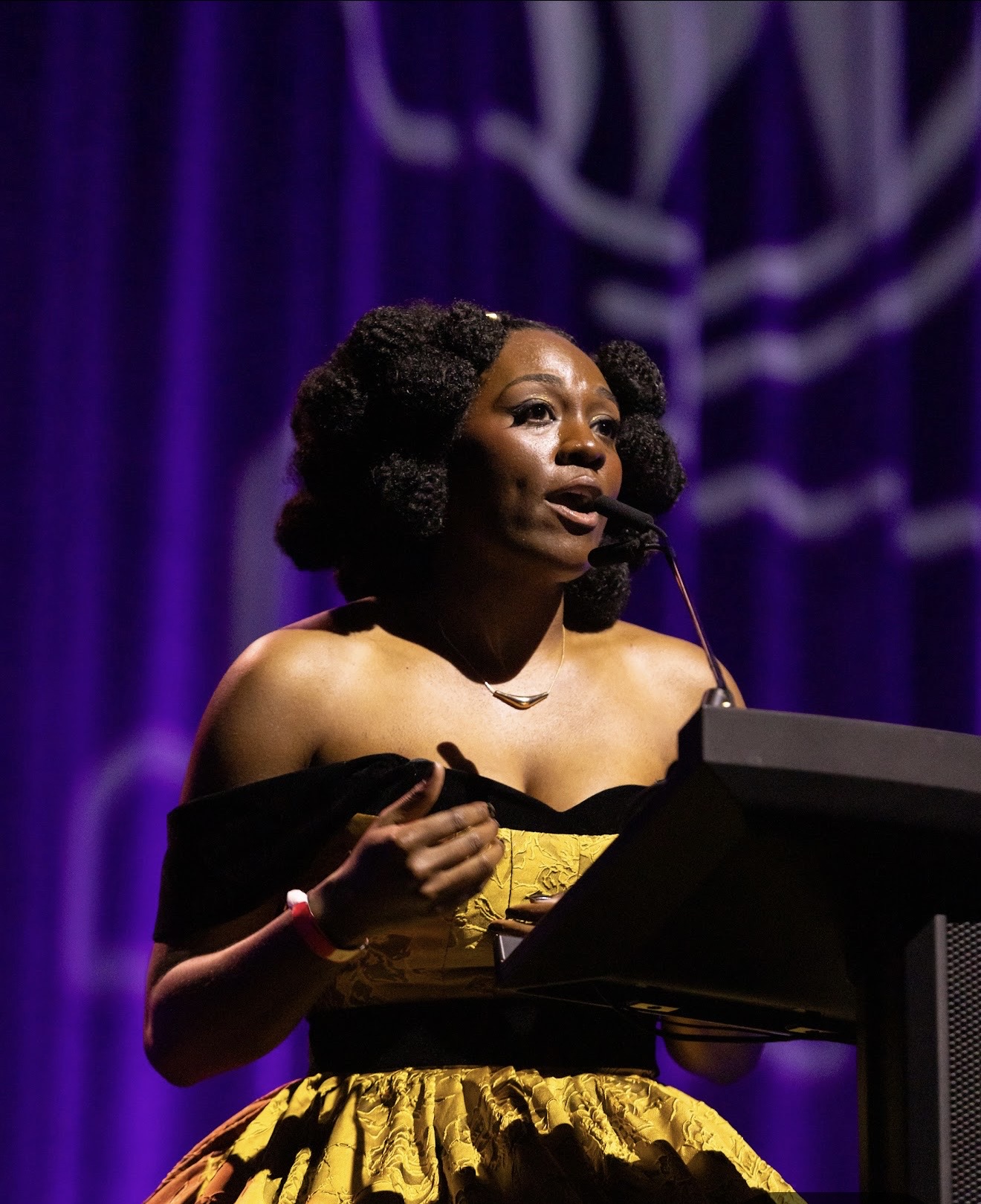 Woman speaking at a podium with purple curtains in the background, wearing a yellow and black off-shoulder dress.