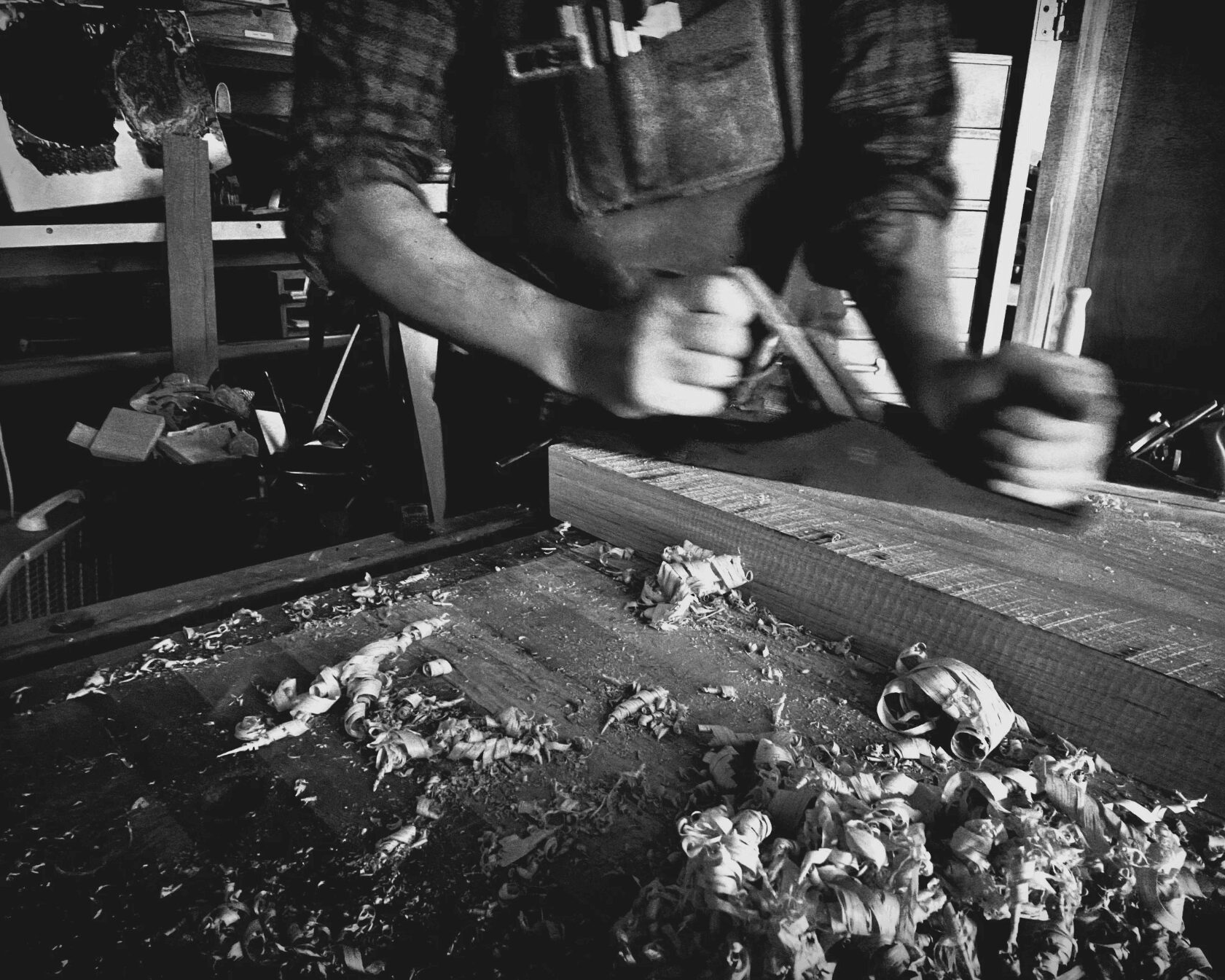 Person working at a cluttered workbench with tools and debris, wearing a vest and plaid shirt, in black and white.
