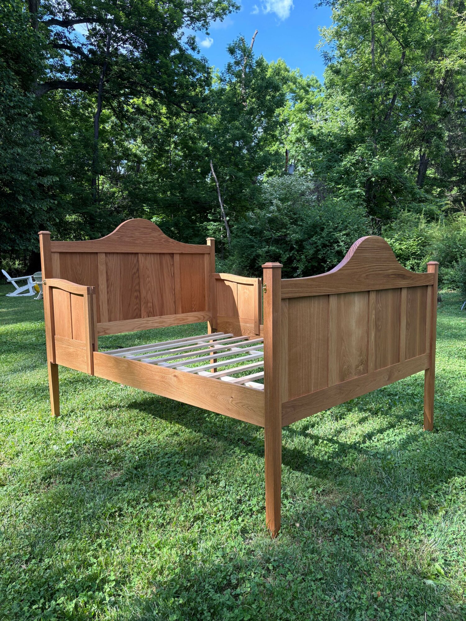 Wooden bed frame outdoors on grass with trees in background, viewed from the side.