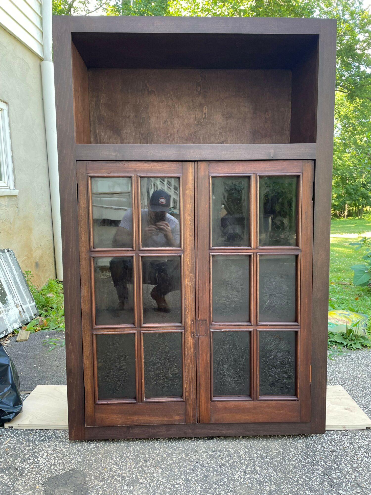 Wooden cabinet with glass-paneled doors, open shelf on top, outdoors with grass and pavement visible