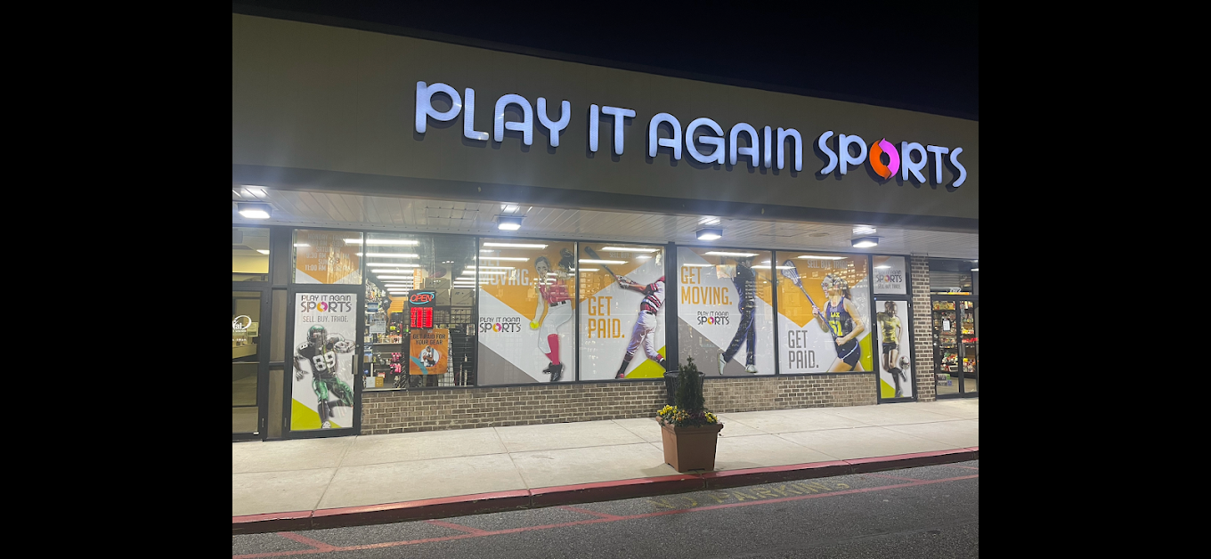 Storefront with sign 'PLAY IT AGAIN SPORTS' and large window displays, night scene, potted plant outside, sidewalk in front.