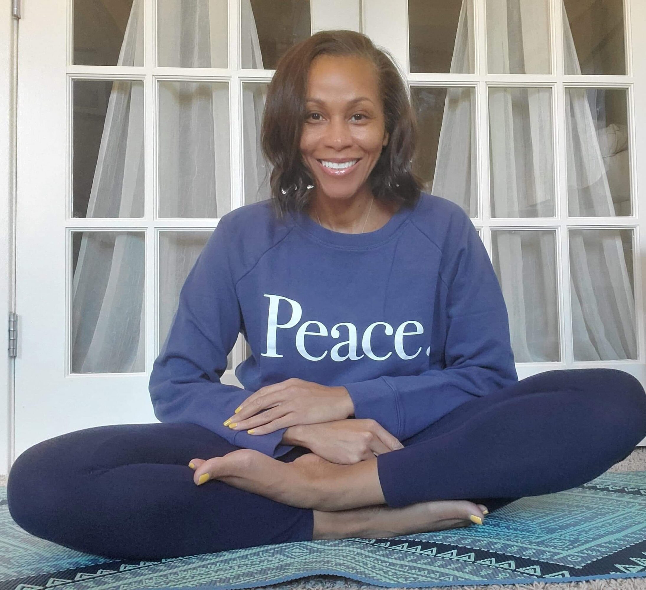 Woman sitting cross-legged on a mat, smiling, wearing a blue sweatshirt with 'Peace' written on it, in front of a window with curtains.
