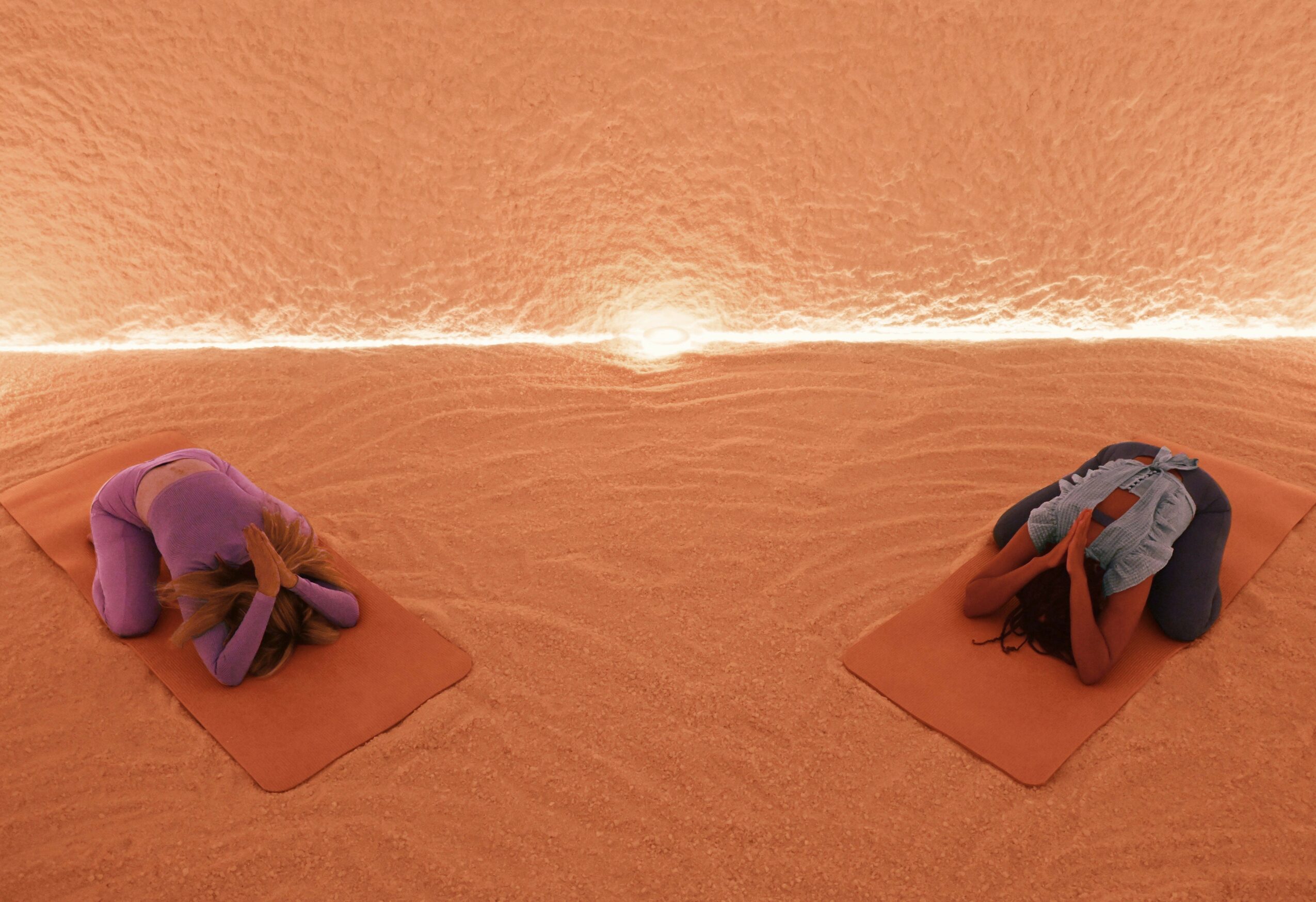 Two people practicing yoga on mats in a desert-like environment with a glowing horizon, facing downward in child's pose.