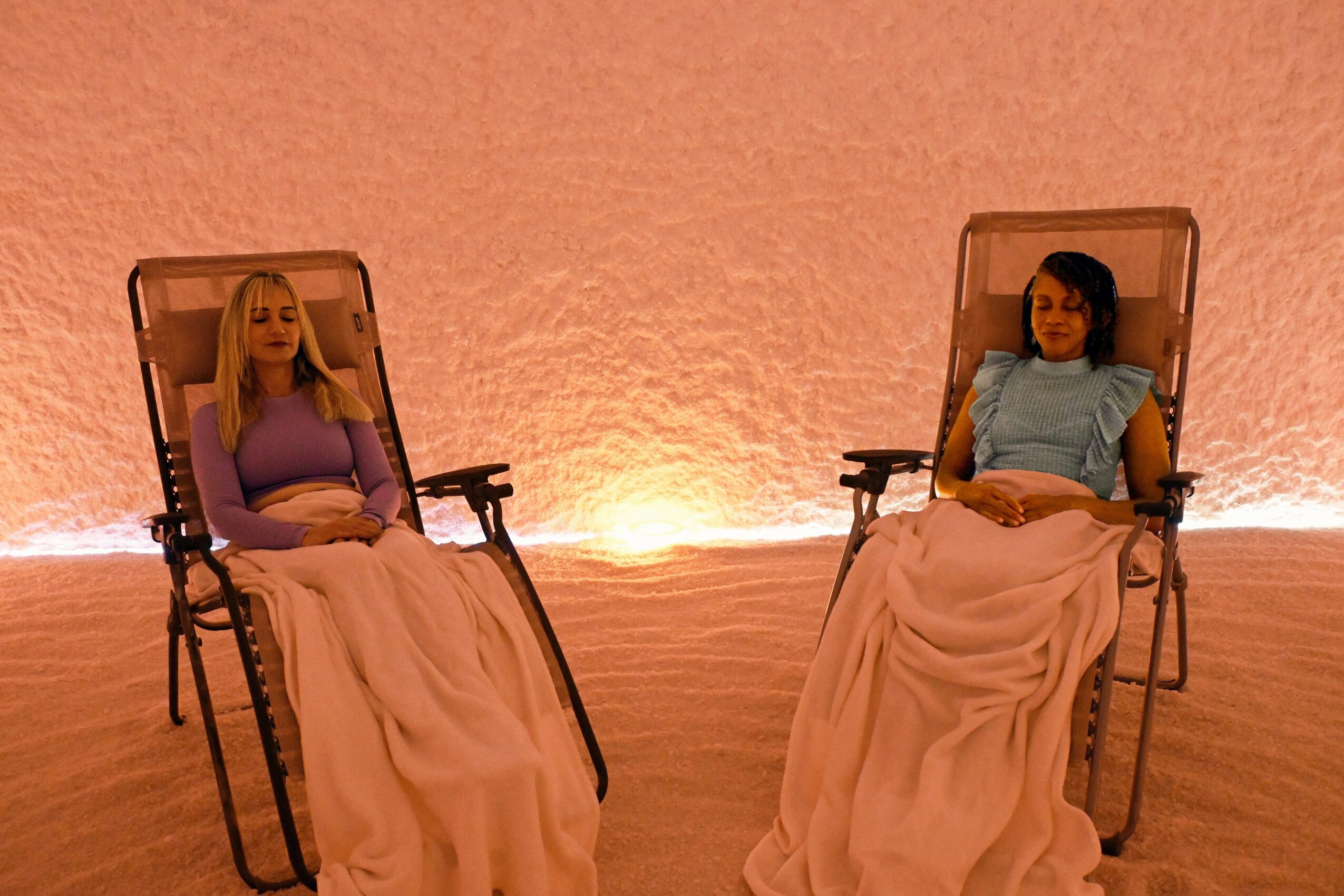Two women sit in reclining chairs with pink blankets, against a pink wall with warm lighting behind them.