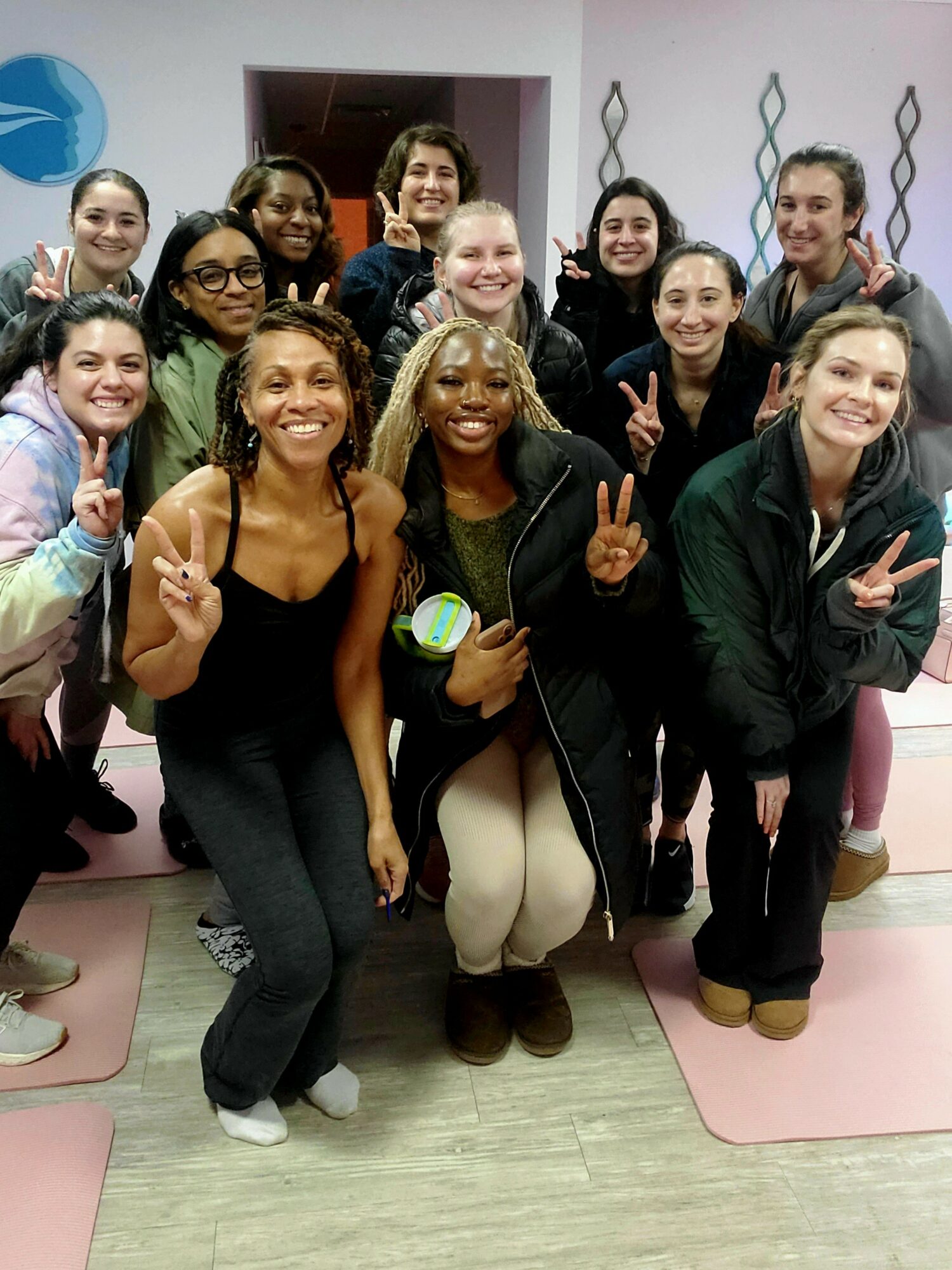 Group of women smiling and making peace signs indoors, some holding yoga mats, in a room with pink walls.