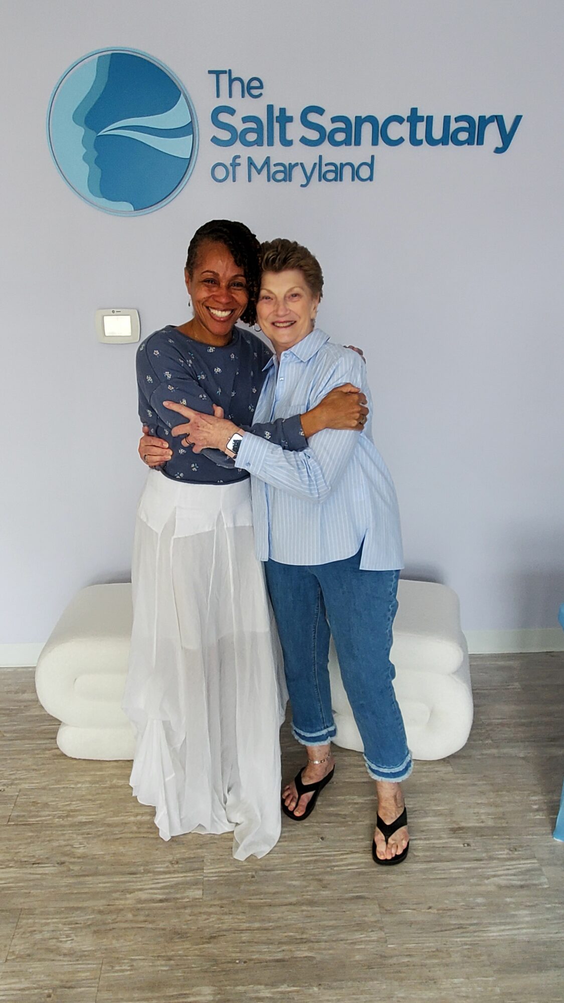 Two women standing together in front of a wall with a logo and text, one in a white dress and the other in a blue top and jeans.
