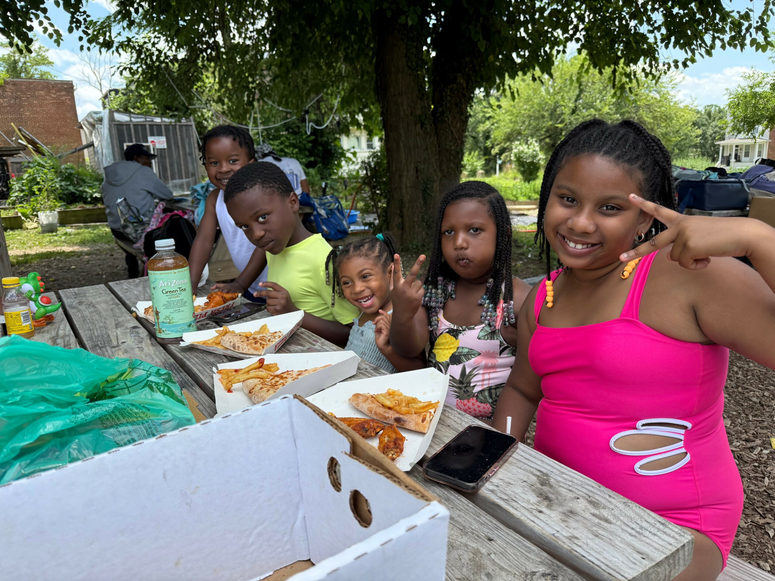 Group of children sitting at a picnic table outdoors, eating pizza, with trees and cars in the background.