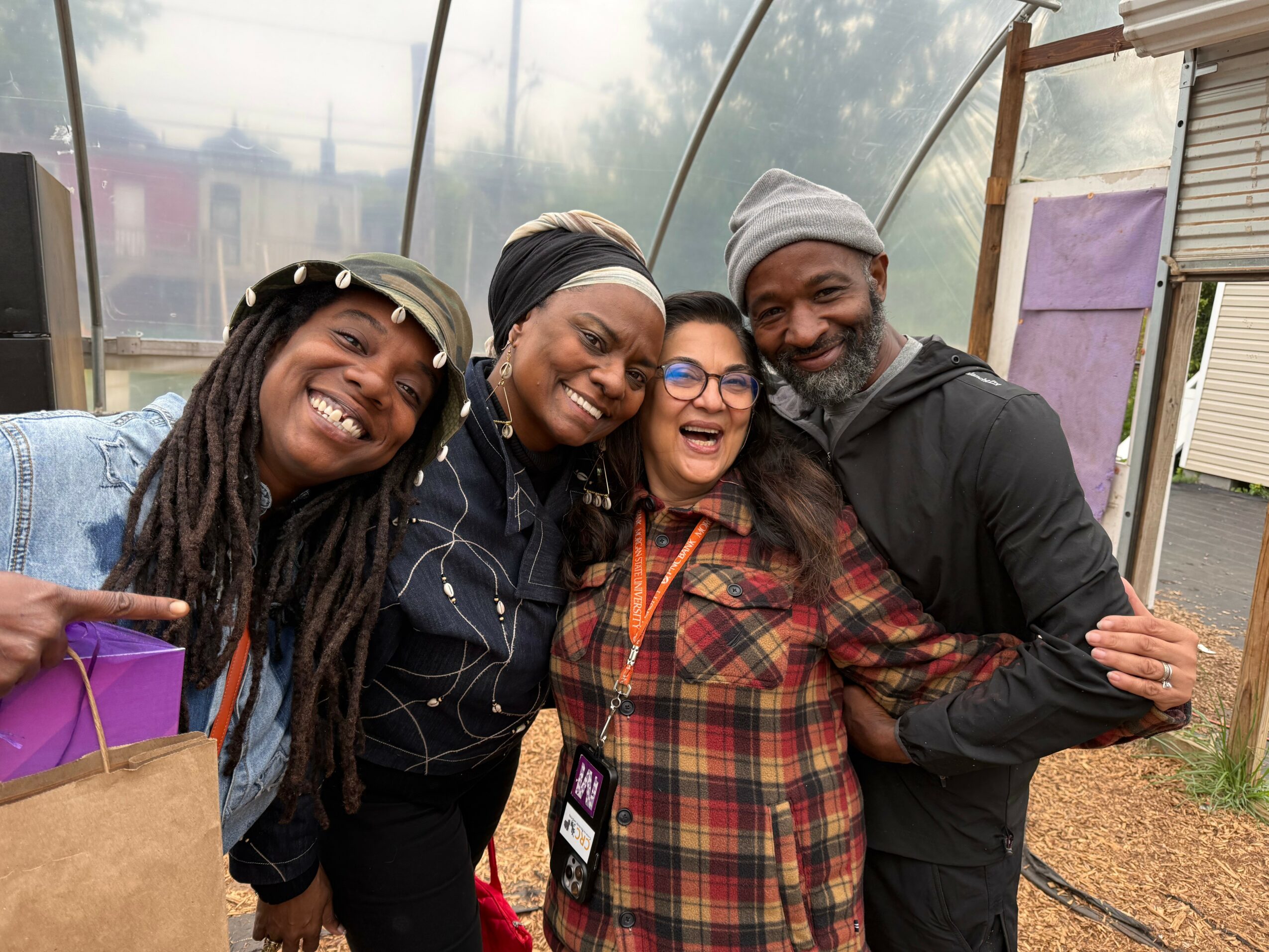 Four people smiling and hugging inside a greenhouse, with a purple bag and a purple towel visible.