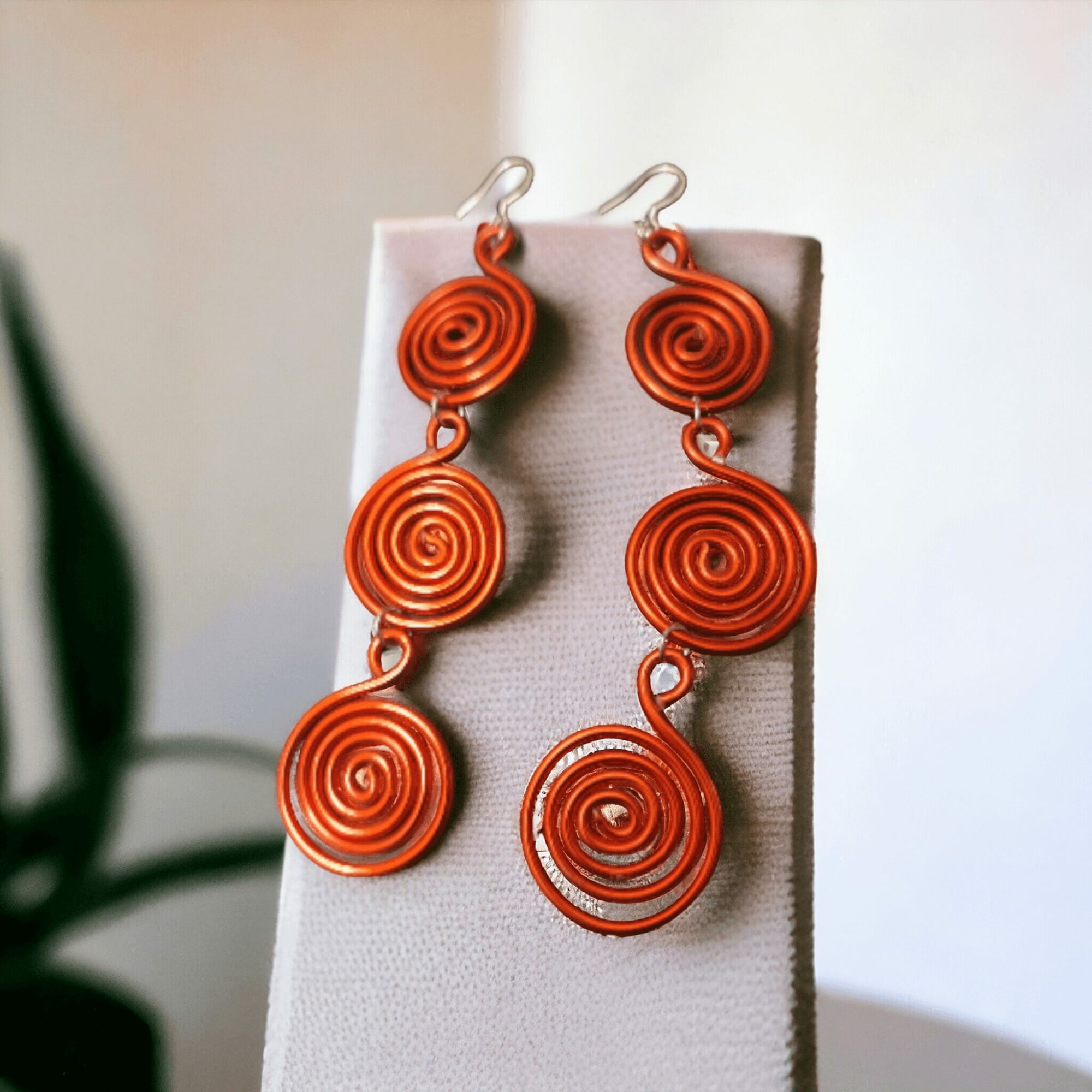 Pair of orange spiral earrings hanging on a display, with a blurred background and a plant partially visible on the left.
