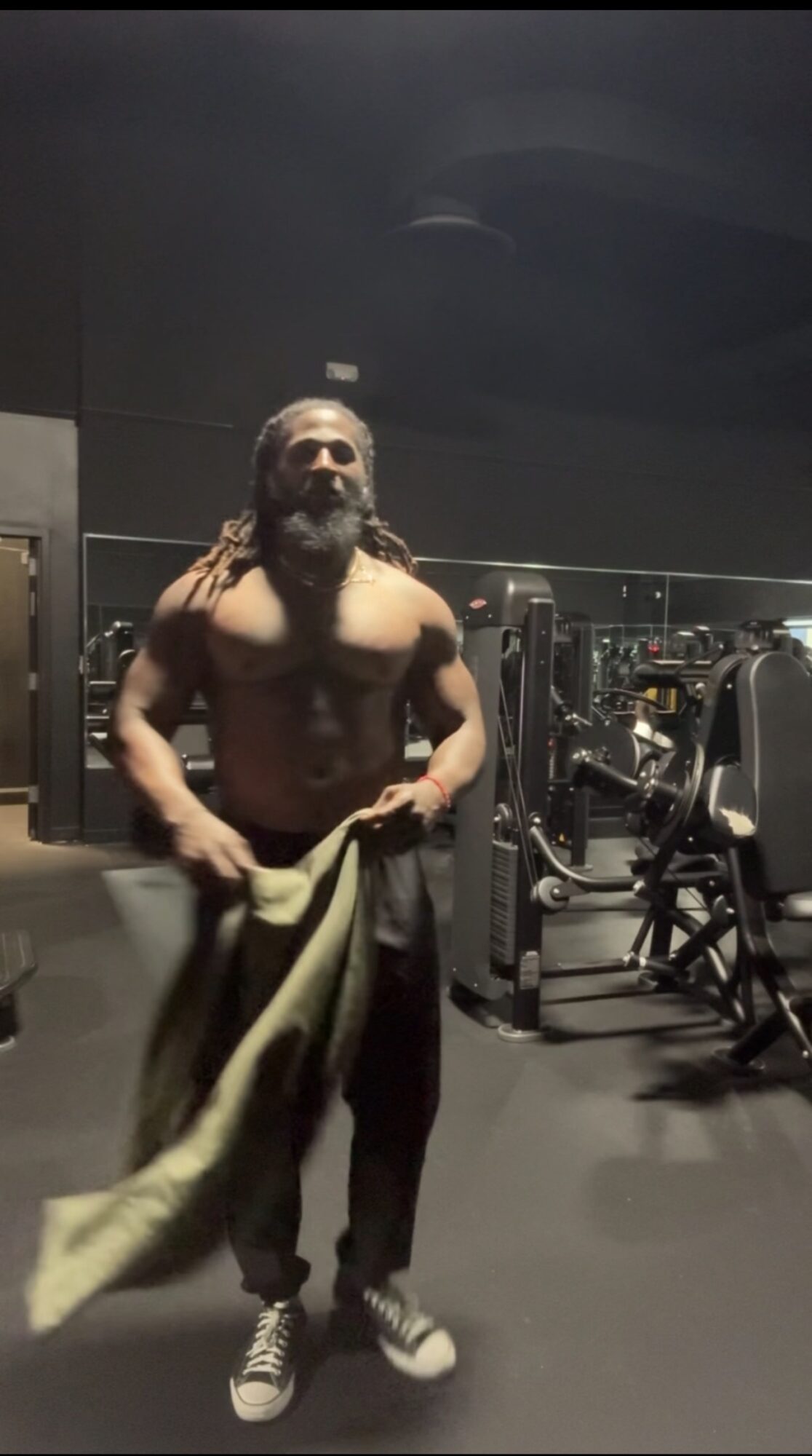 A shirtless man with a beard and long hair stands in a gym, holding a towel, with gym equipment in the background.