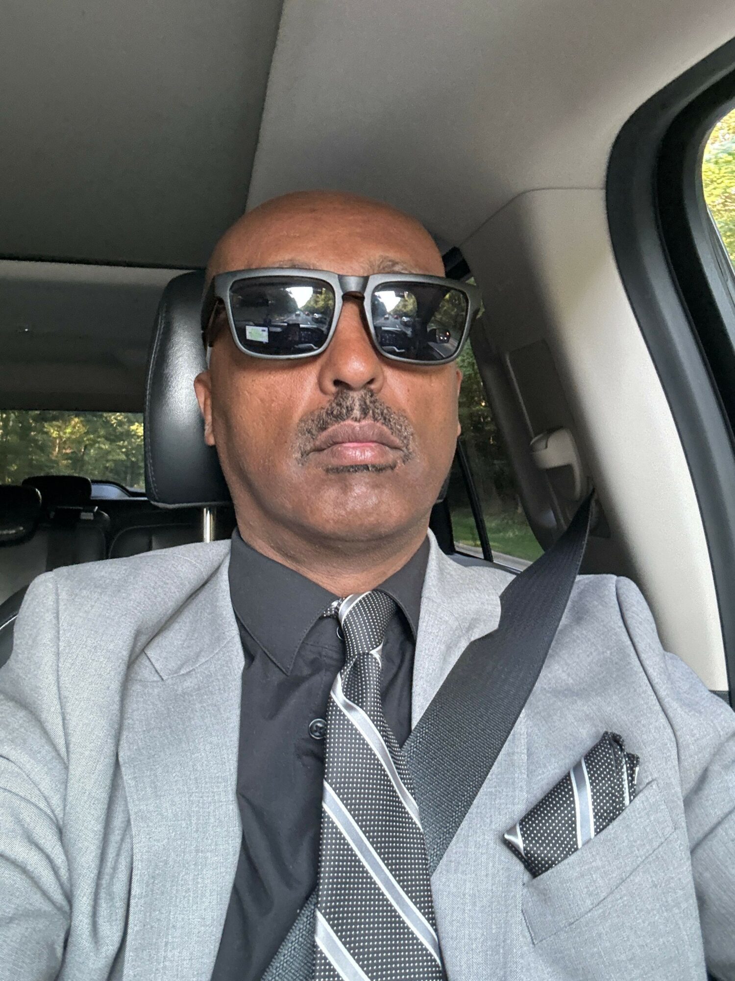Man wearing sunglasses, gray suit, and tie, sitting in a vehicle with seatbelt on, window and greenery visible outside.