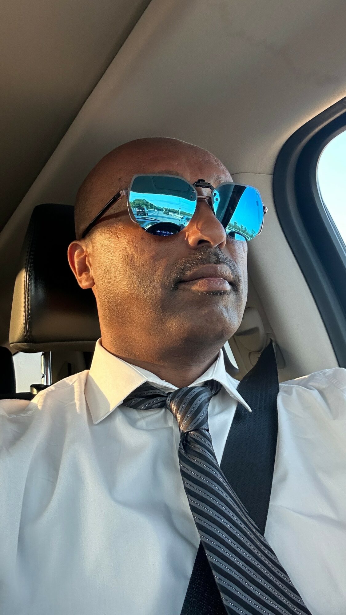 Man wearing sunglasses and a white shirt with a striped tie, sitting in a vehicle, looking out the window.