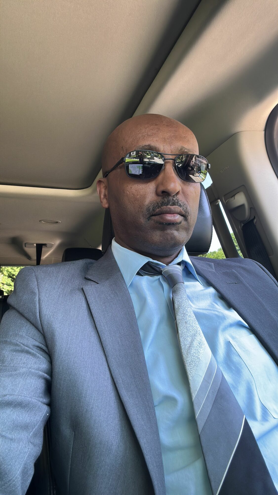 Man wearing sunglasses and a suit, sitting in a vehicle, looking at the camera.
