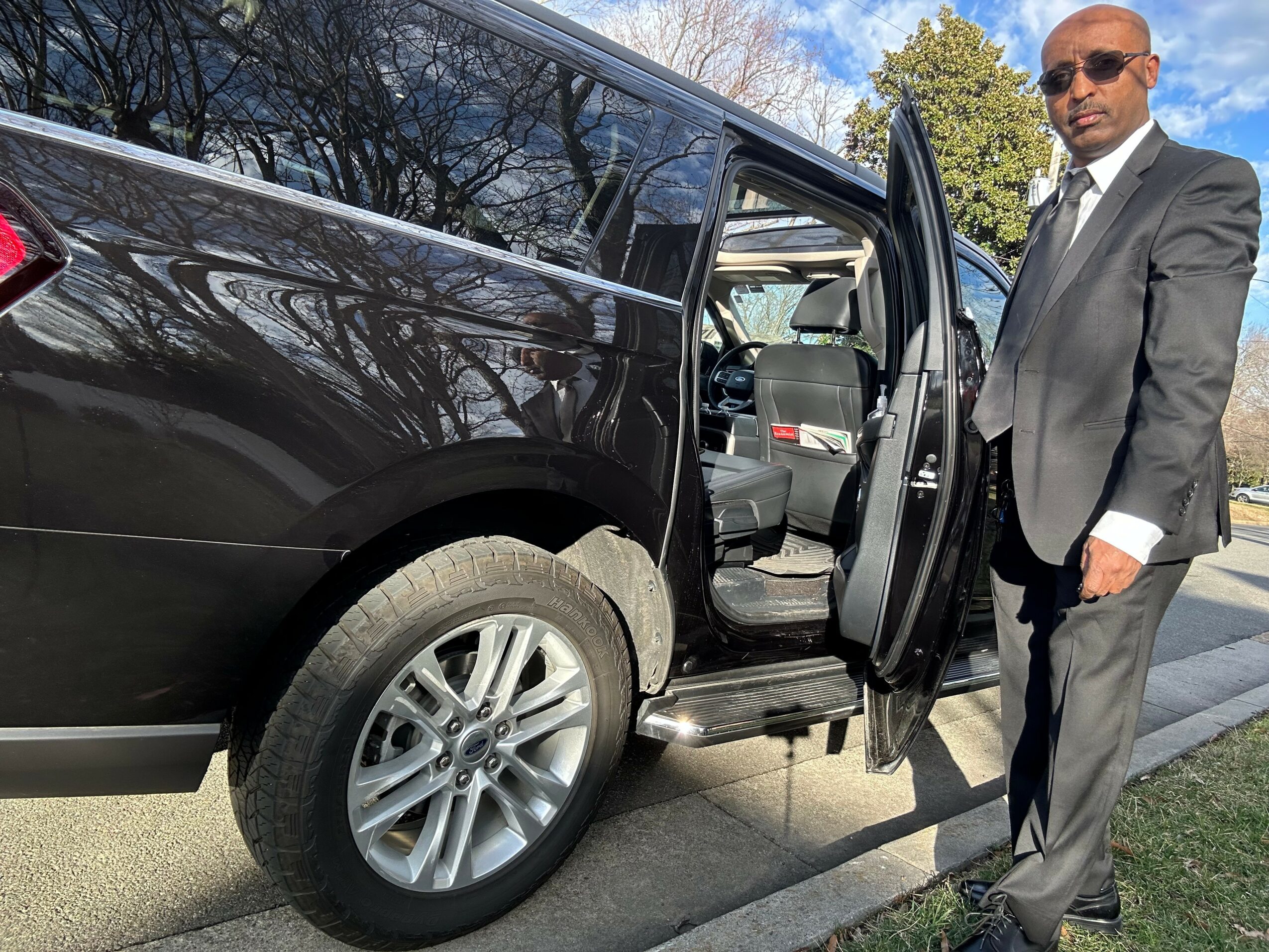 Man in suit standing outside a black SUV with open door, parked on a street with trees and blue sky.