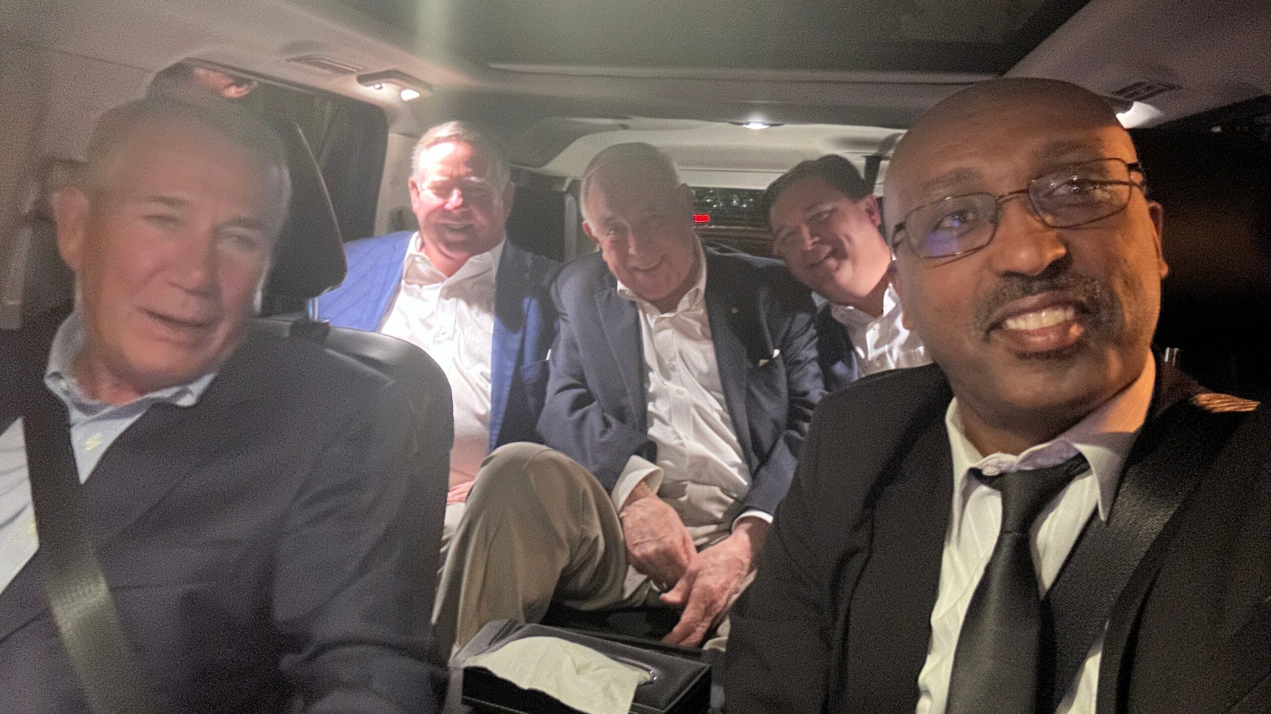 Five men sitting in a vehicle, some looking at the camera, with a dark interior background.