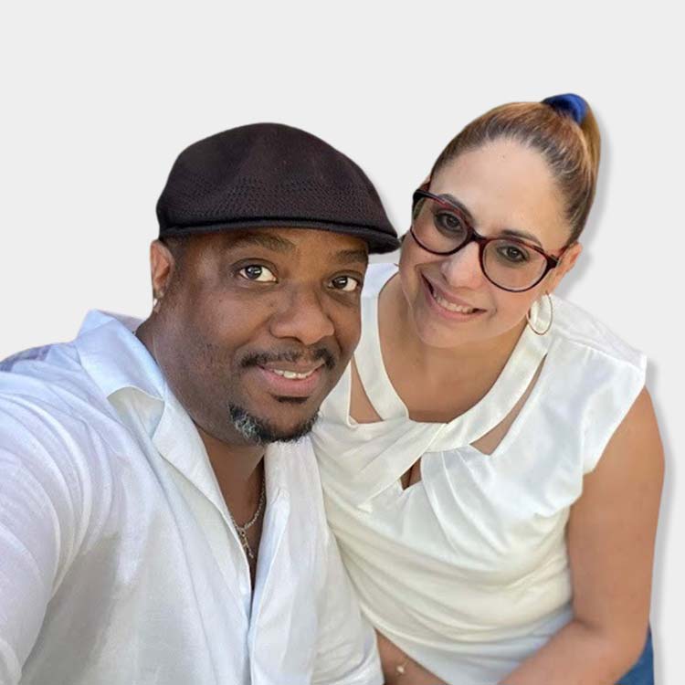 Two people smiling, one wearing a black cap and white shirt, the other with glasses and a white top, against a white background.