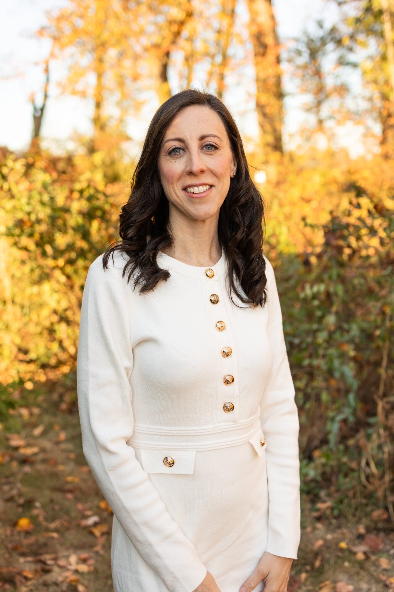 Woman with dark hair in a white outfit outdoors with autumn trees in background.