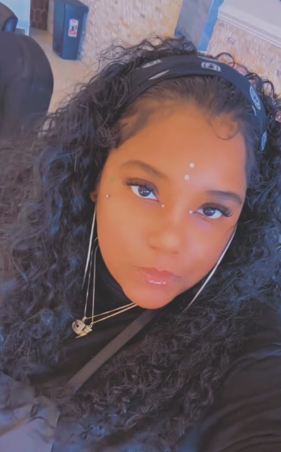 Young woman with curly hair, wearing headphones and jewelry, looking at the camera with a neutral expression.
