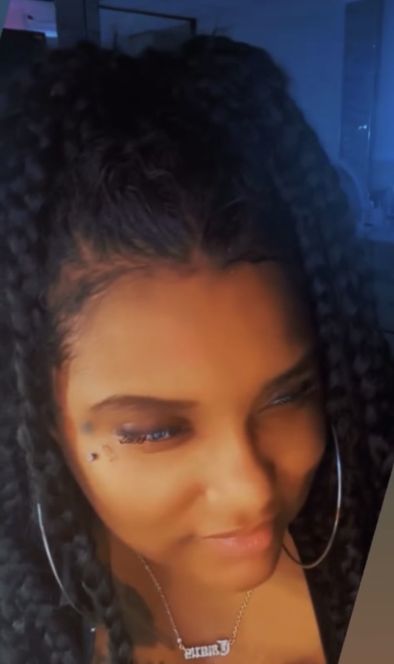 Close-up of a woman with curly hair, hoop earrings, and a necklace, smiling slightly.