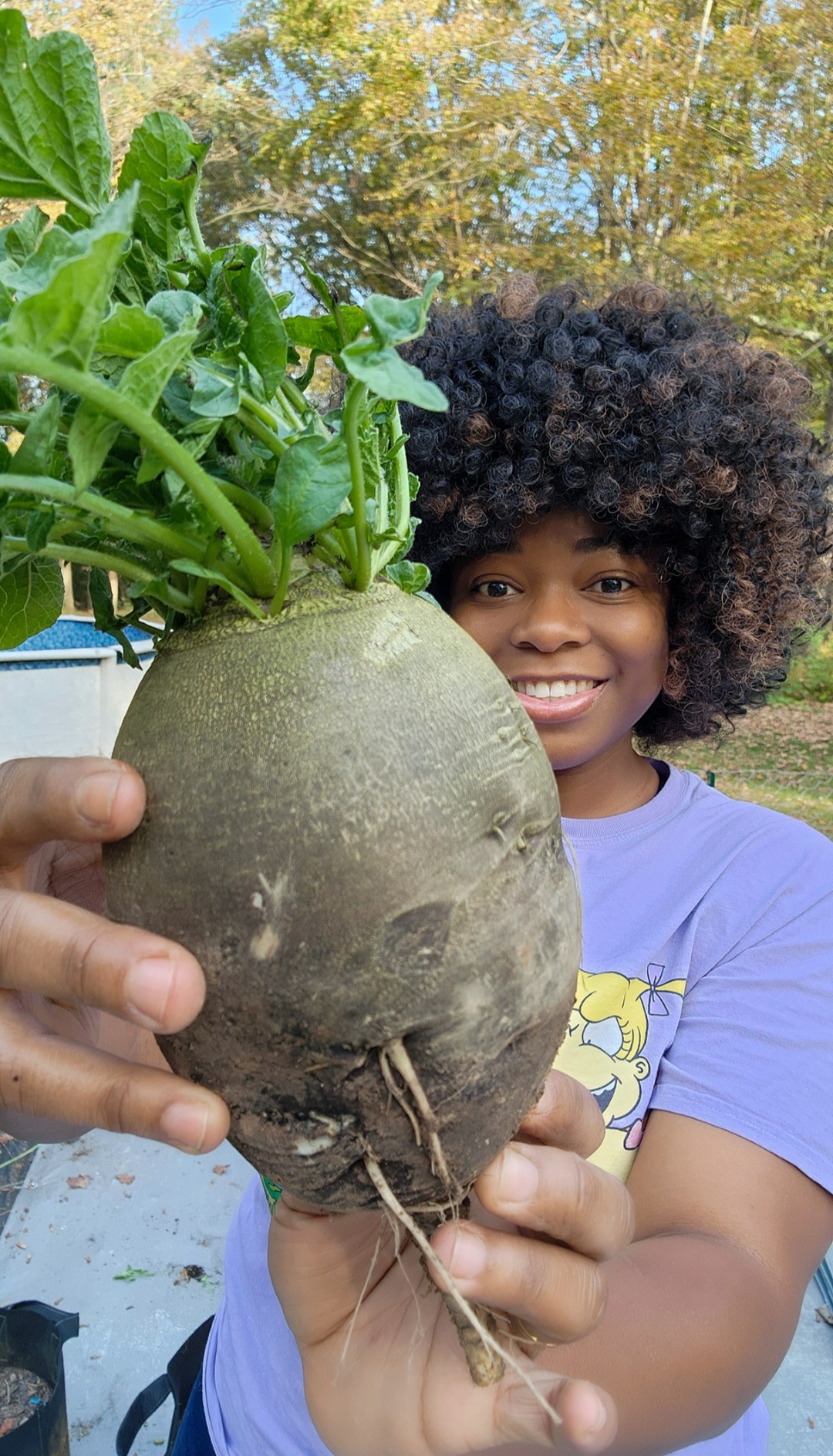 Child with curly hair holding a large radish with green leaves outdoors, smiling, trees in background.