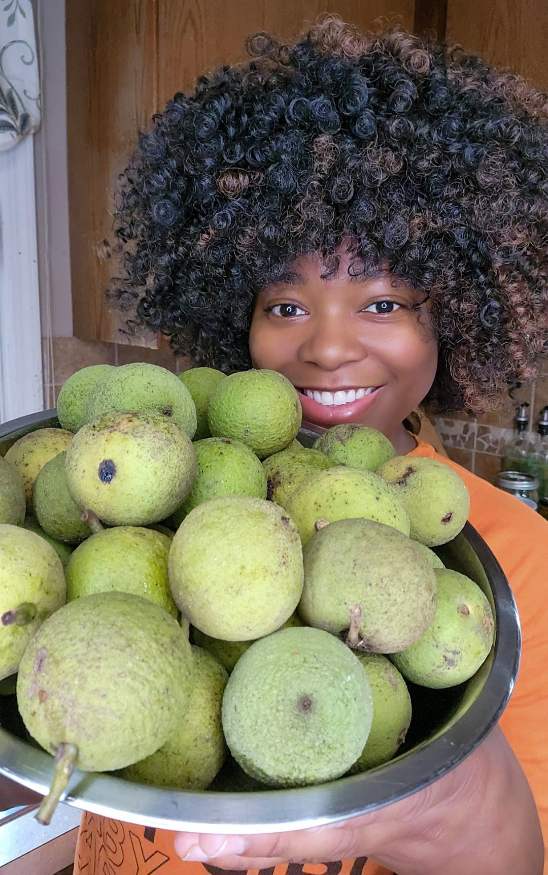 Person with curly hair smiling behind a large bowl of green pears, holding the bowl with both hands.