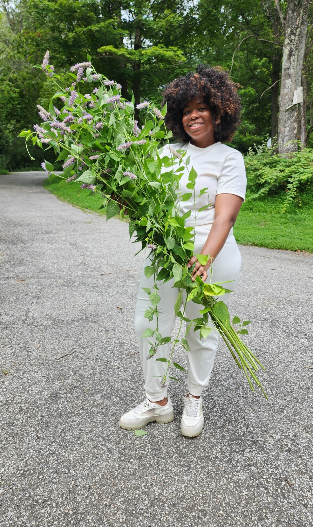 Person with curly hair smiling, holding a large bouquet of flowers outdoors on a gravel path.