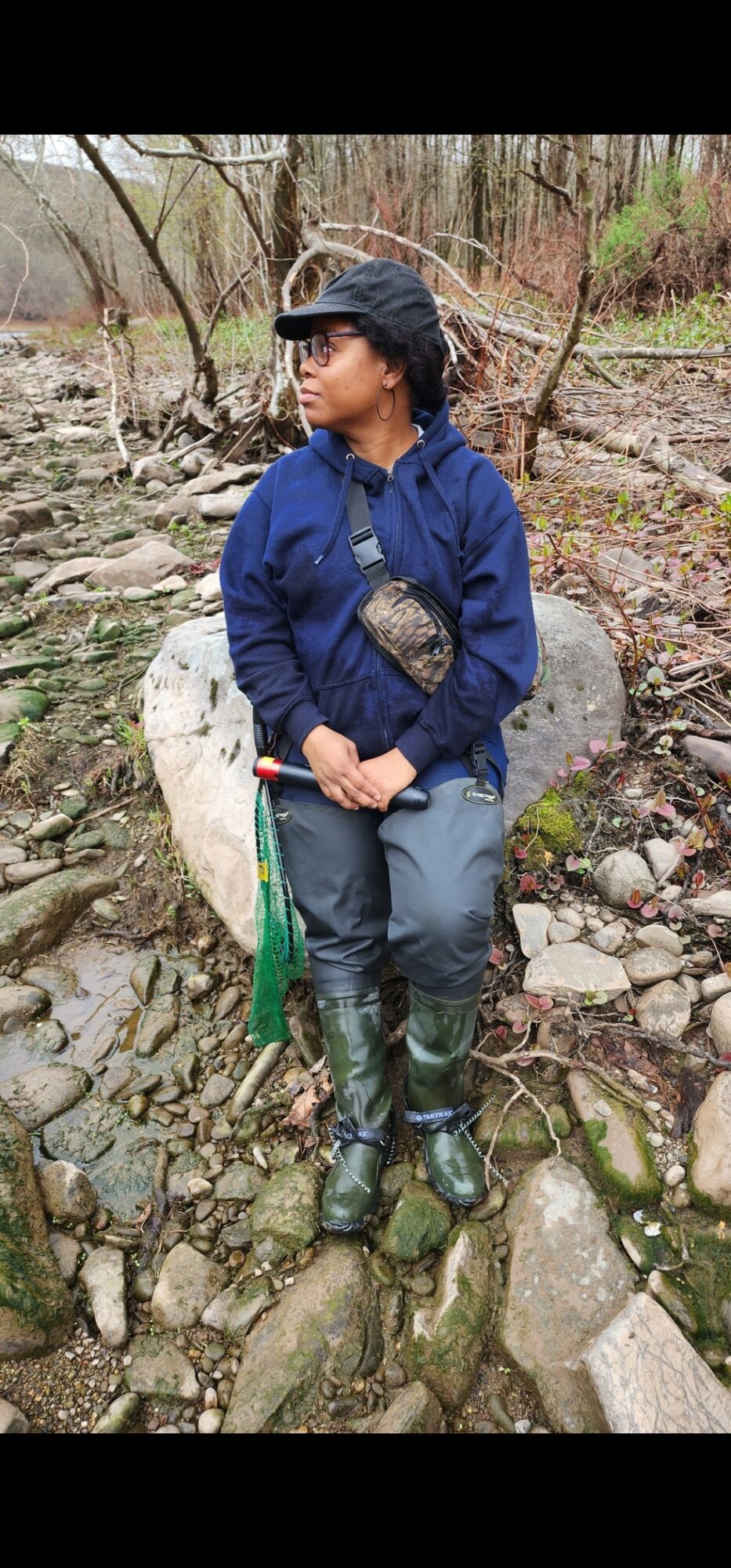 Person sitting on rocks in a wooded area, wearing a blue jacket, cap, and sunglasses, holding a walking stick.