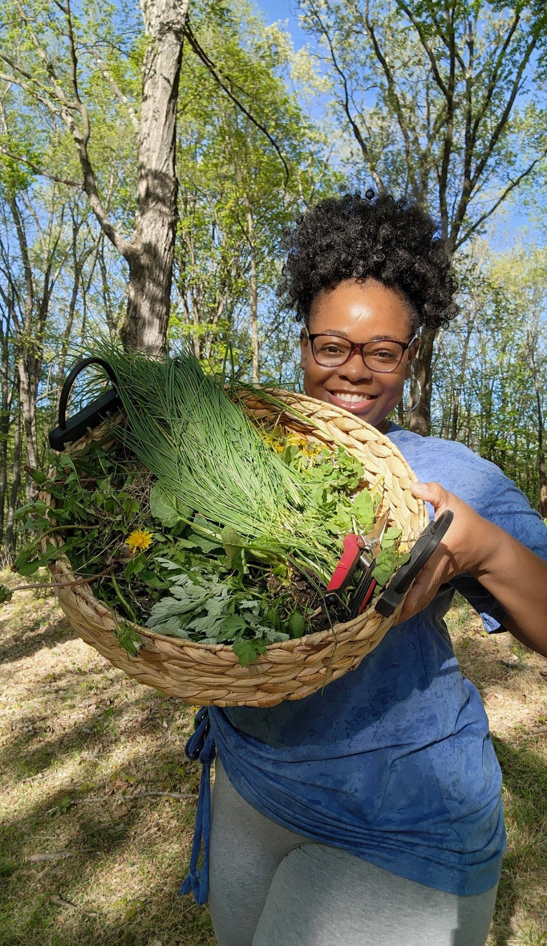 Woman outdoors holding a basket of greenery and flowers, smiling, trees and blue sky in background.