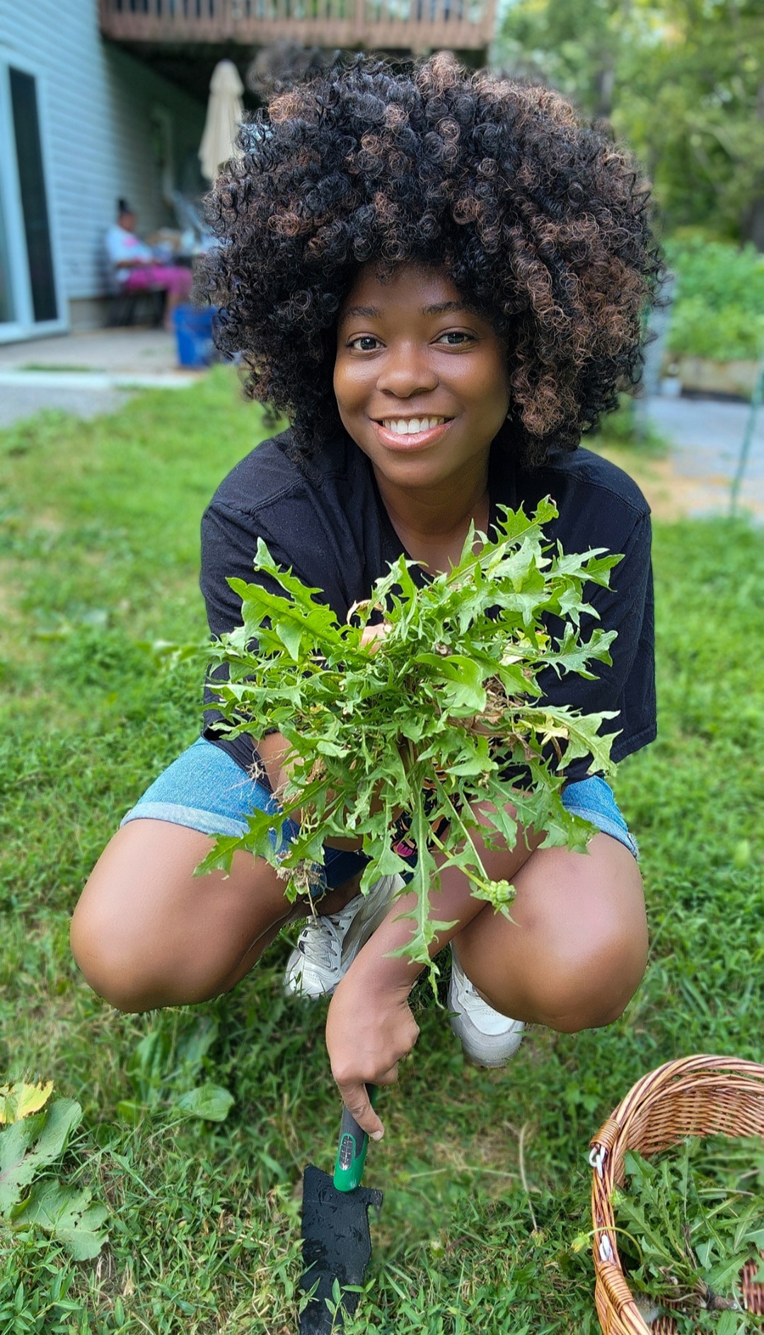 Young woman crouching outdoors holding a leafy plant, smiling, with a garden and house in background.