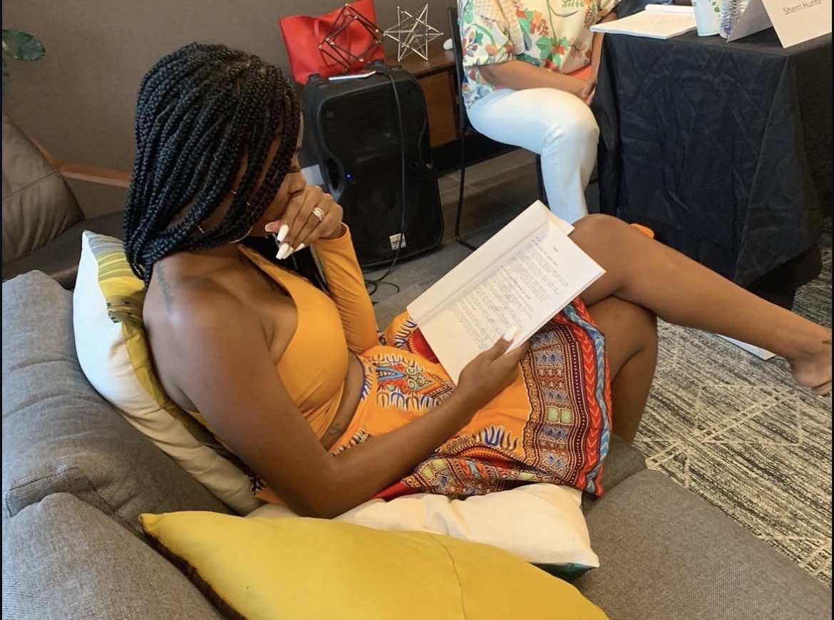 Woman with braided hair reading a book while sitting on a couch with a yellow pillow.