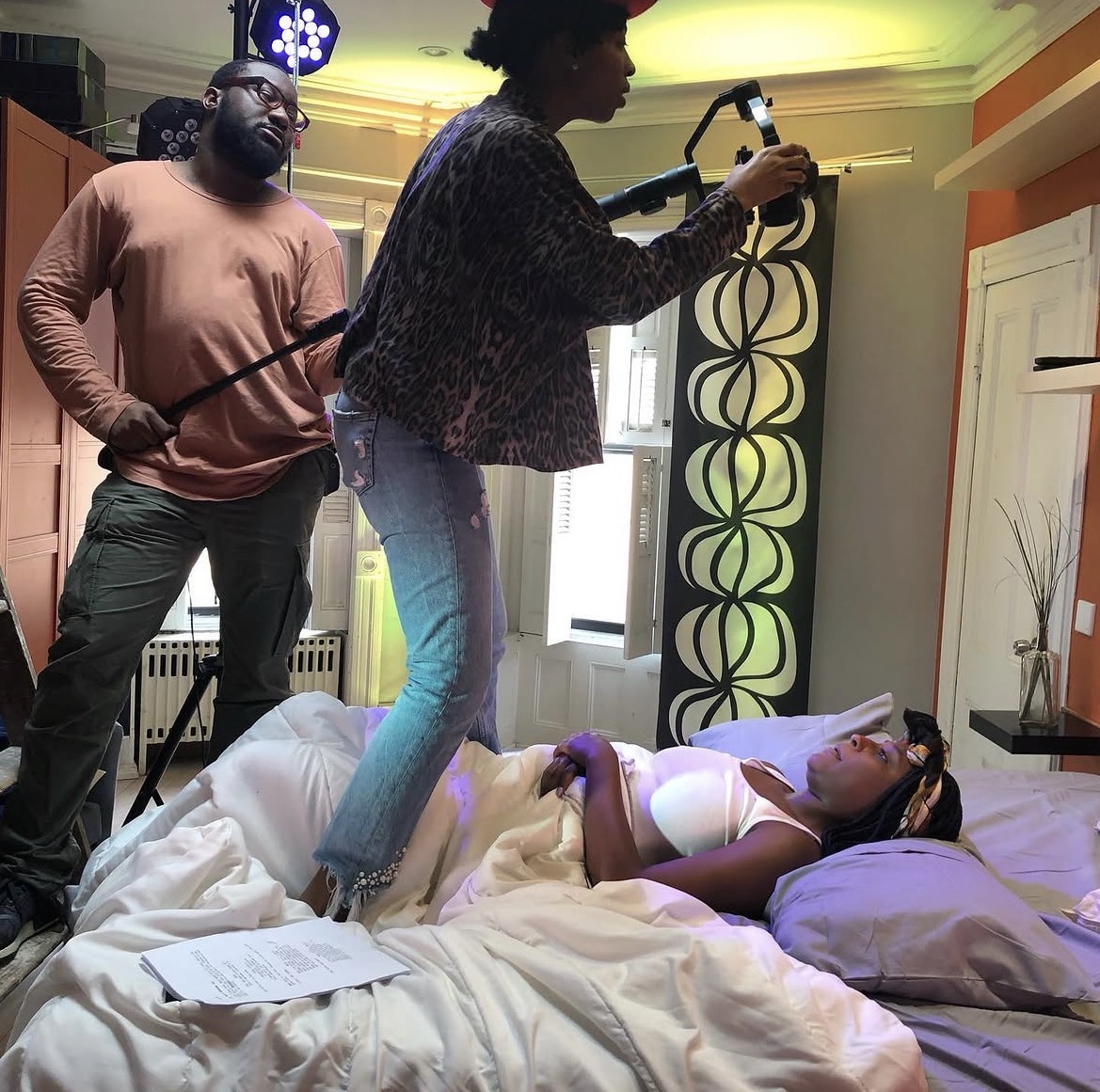 A woman lying on a bed with a woman standing over her, holding a device, while a man observes in a room with colorful decor.