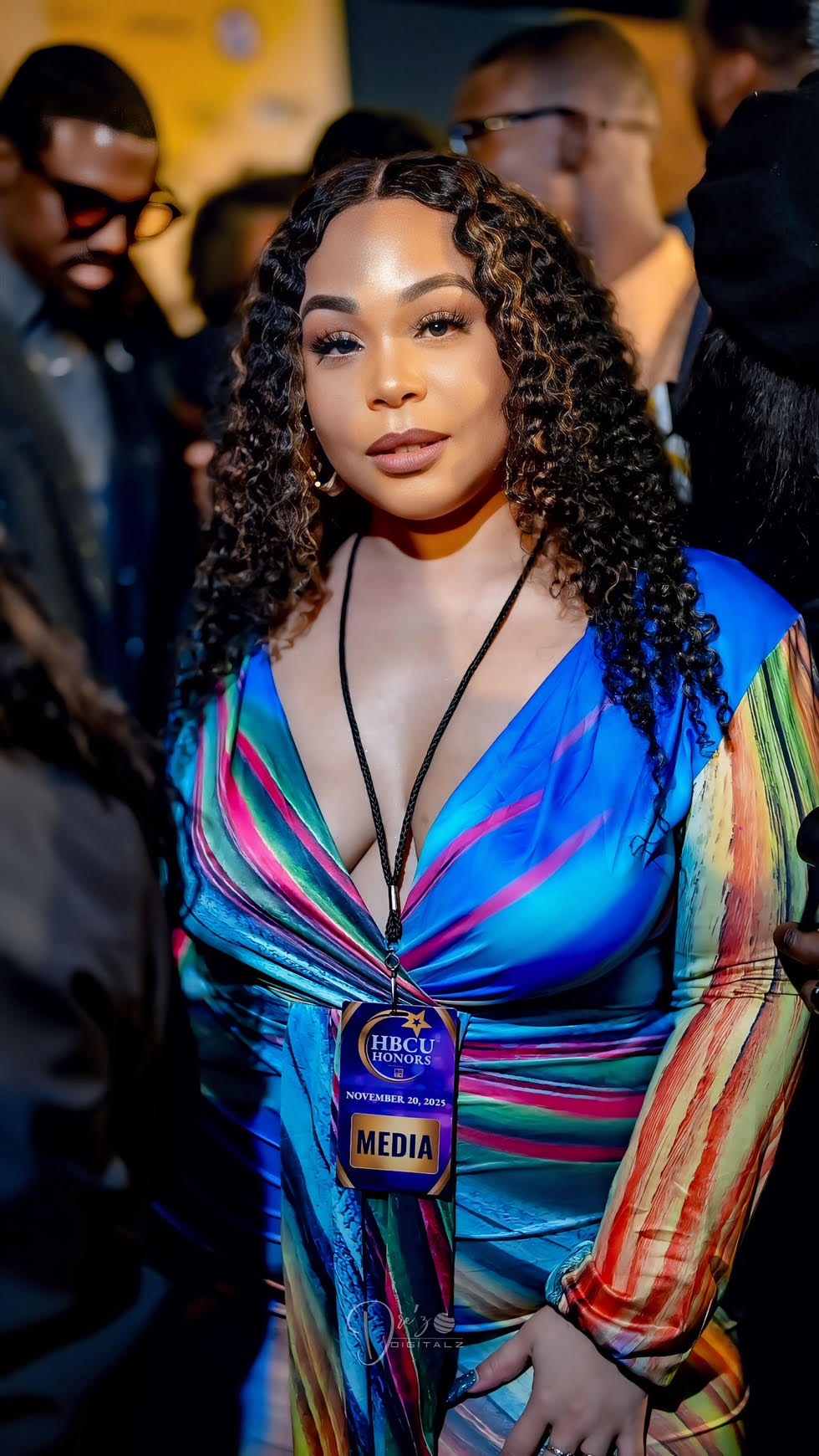 Woman with curly hair wearing a colorful dress and media badge, surrounded by people at an event.