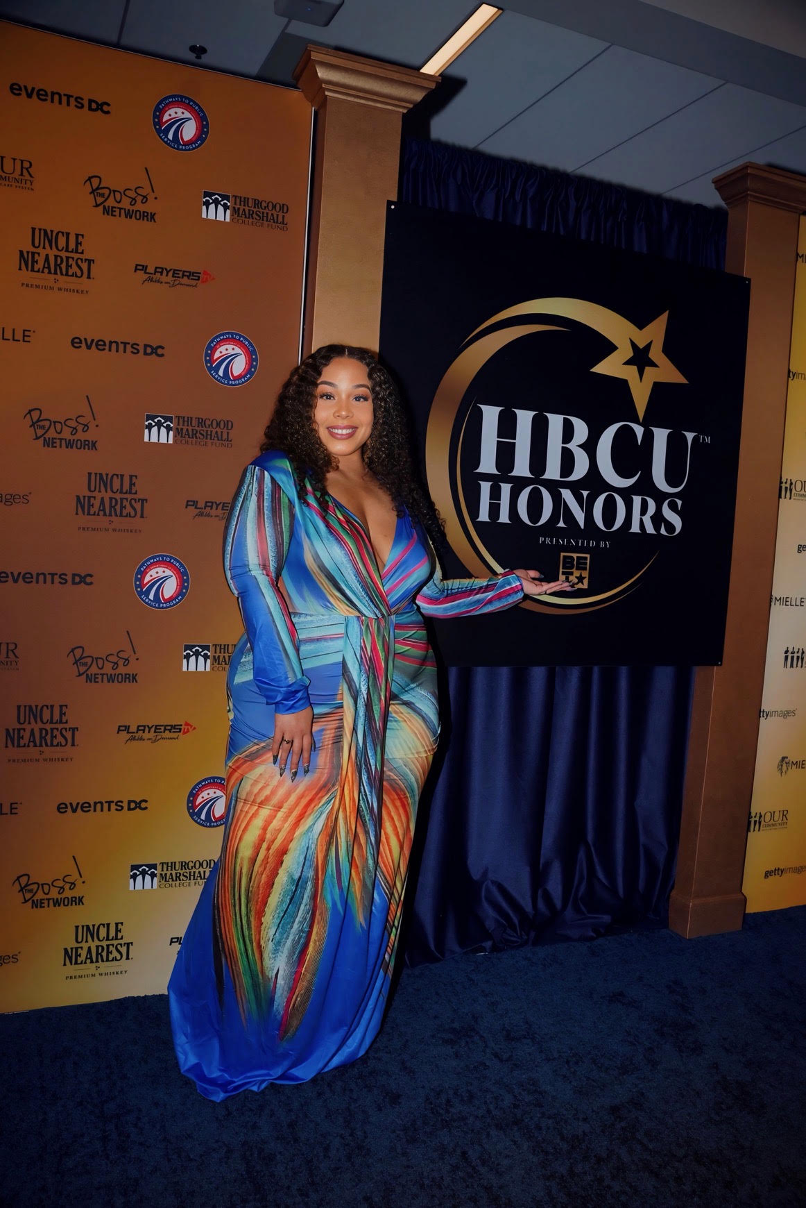 Woman in colorful dress holding award at HBCU Honors event, standing next to a large logo backdrop.