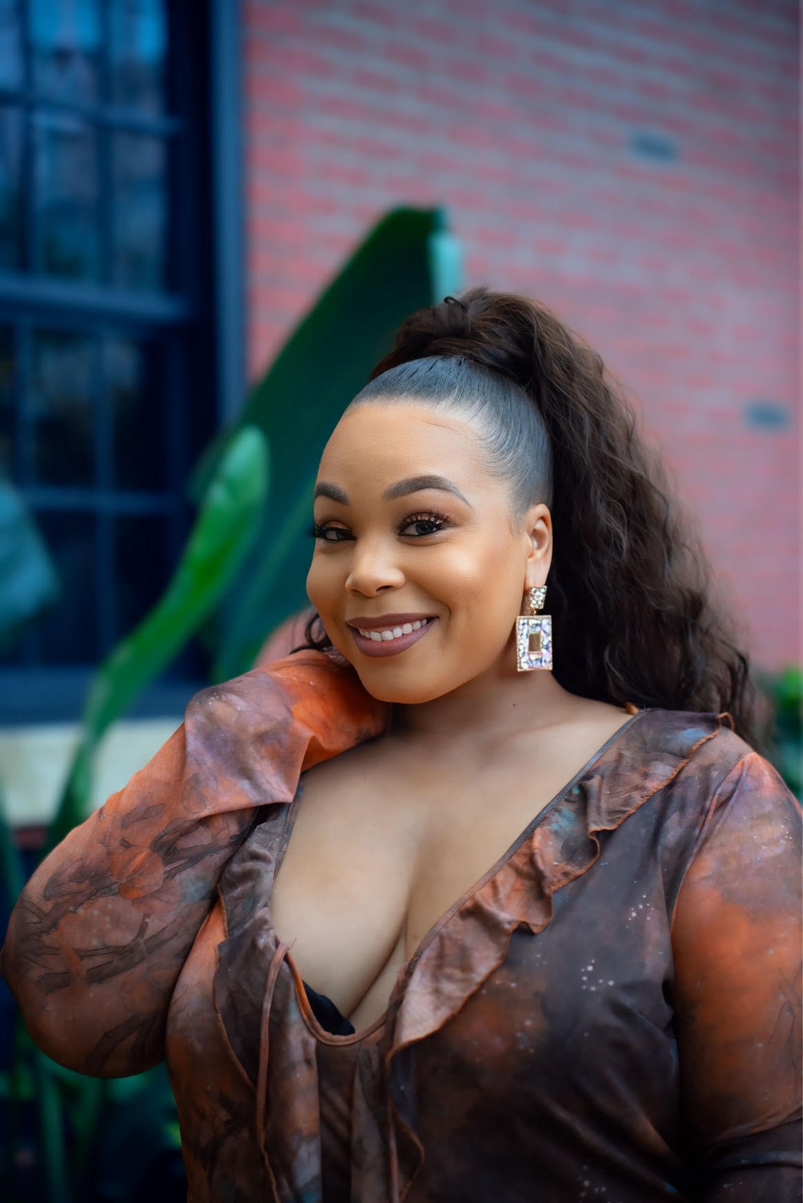 Smiling woman with curly hair in a high ponytail, wearing earrings and a patterned top, outdoors near a brick building.