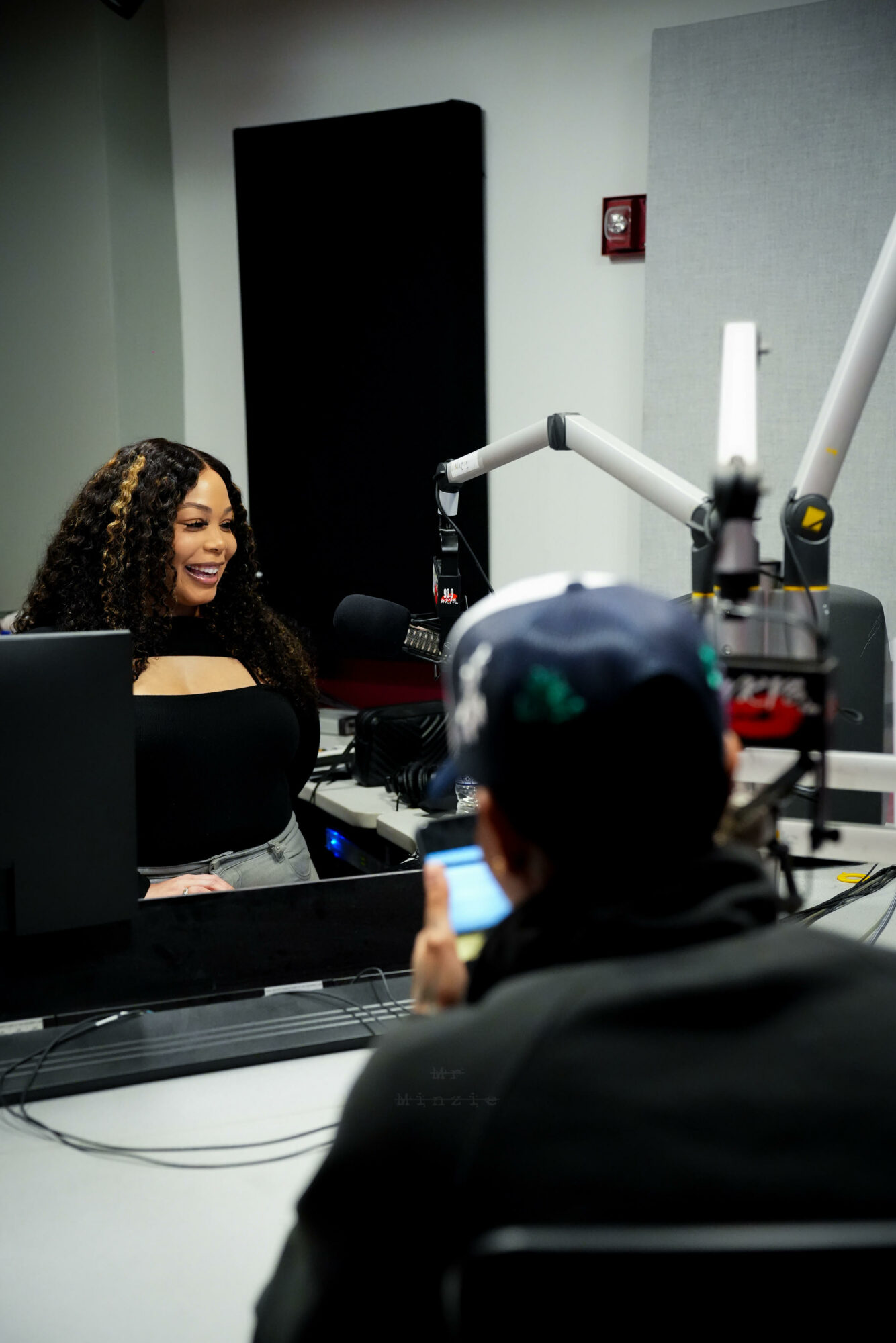 Two people in a radio studio, one woman smiling and speaking into a microphone, the other person using a phone.