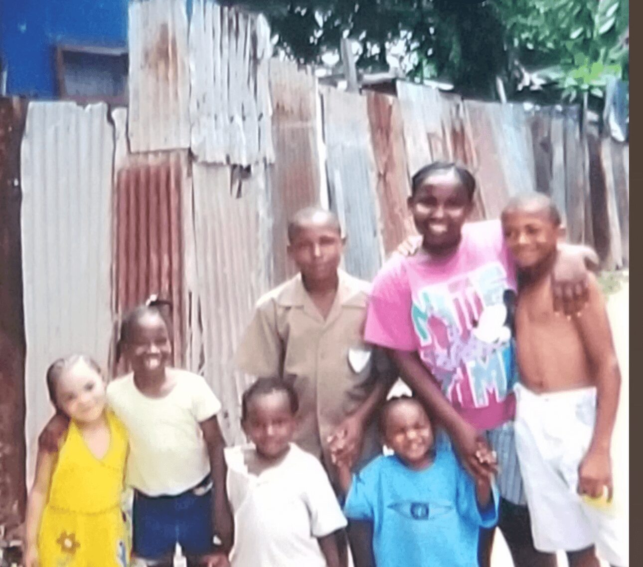 Group of six children standing outdoors in front of a corrugated metal fence, smiling and holding hands.