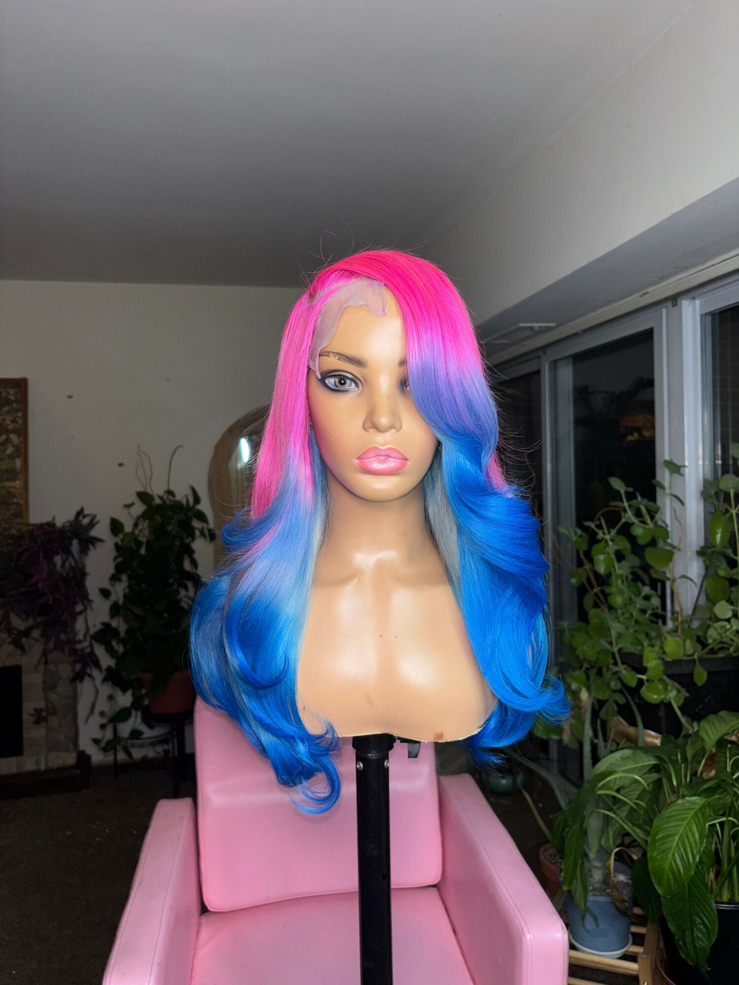 Bust of a woman with colorful pink and blue hair, sitting on a pink chair in a room with plants.